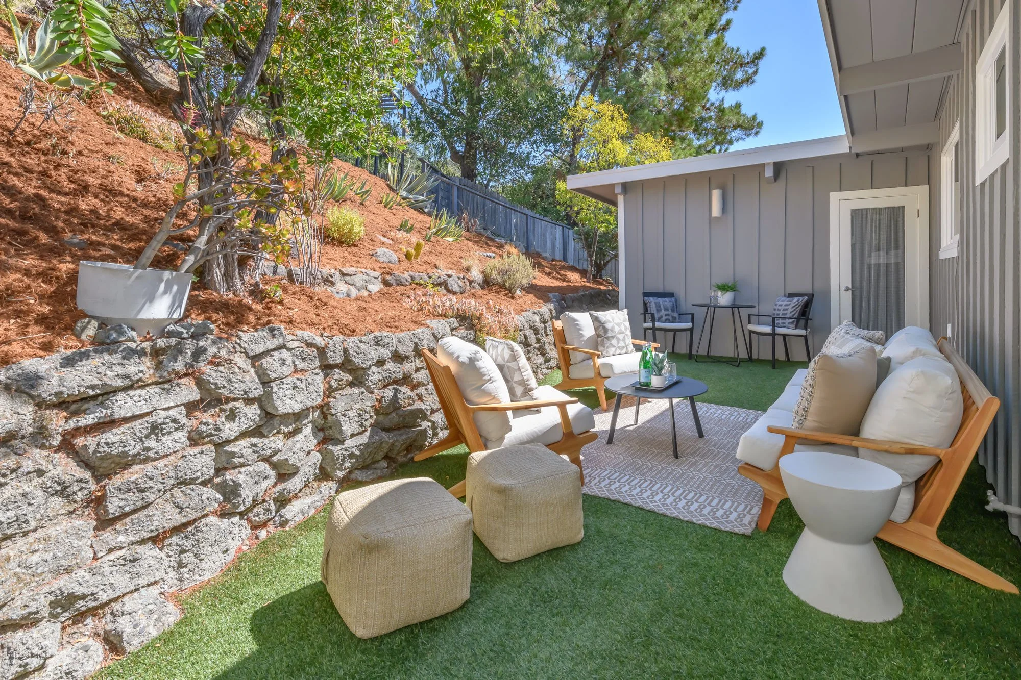 A cozy outdoor patio area with beige and wooden furniture including cushioned chairs and a sofa, a small round table with bottles and glasses, situated on artificial grass, adjacent to a landscaped hillside with plants and volcanic rock, and a house 
