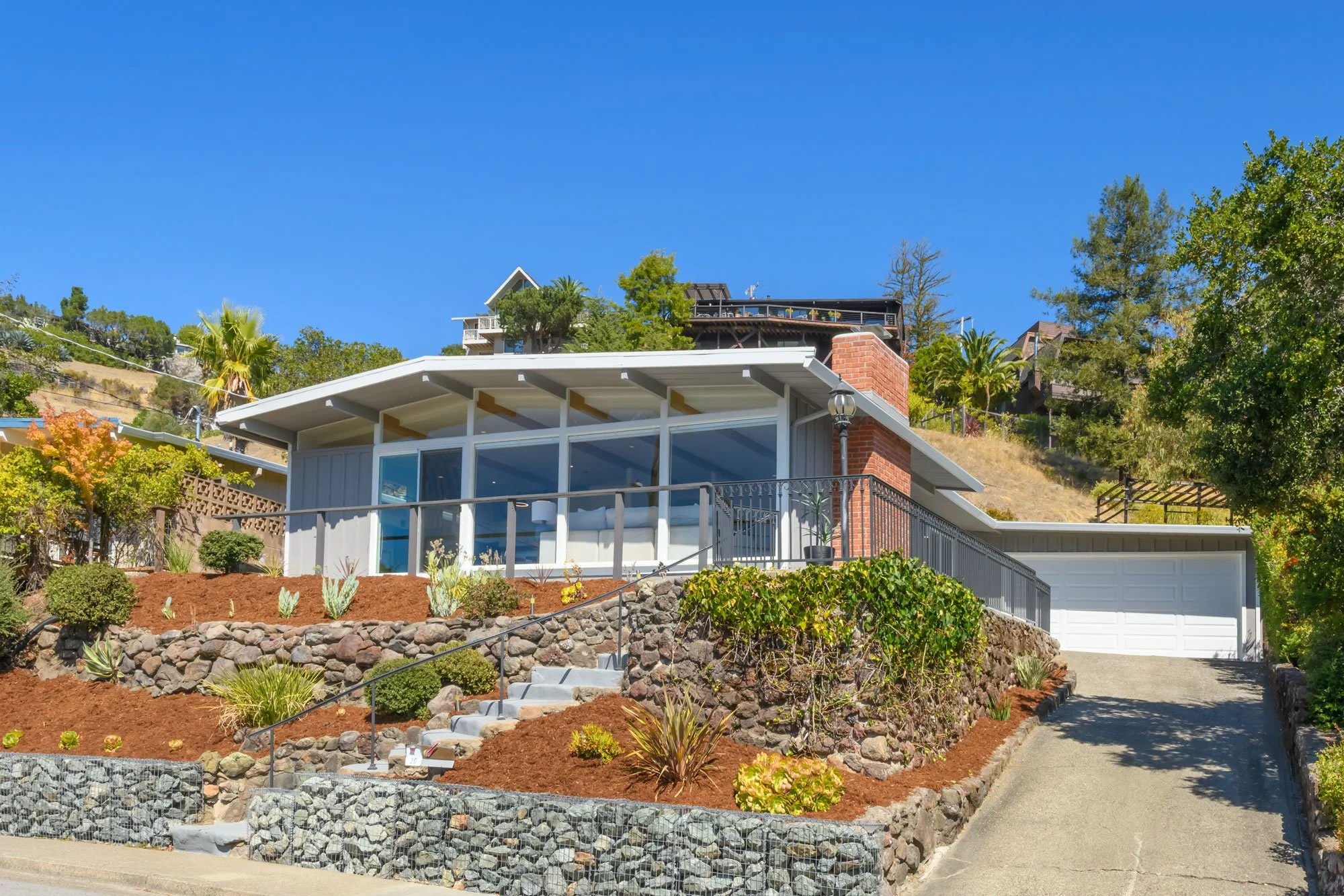 View of a modern house on a hillside with a glass sunroom, landscaped front yard, plants, stone wall, and a driveway leading to a garage, with clear blue sky and neighboring houses on the hill.