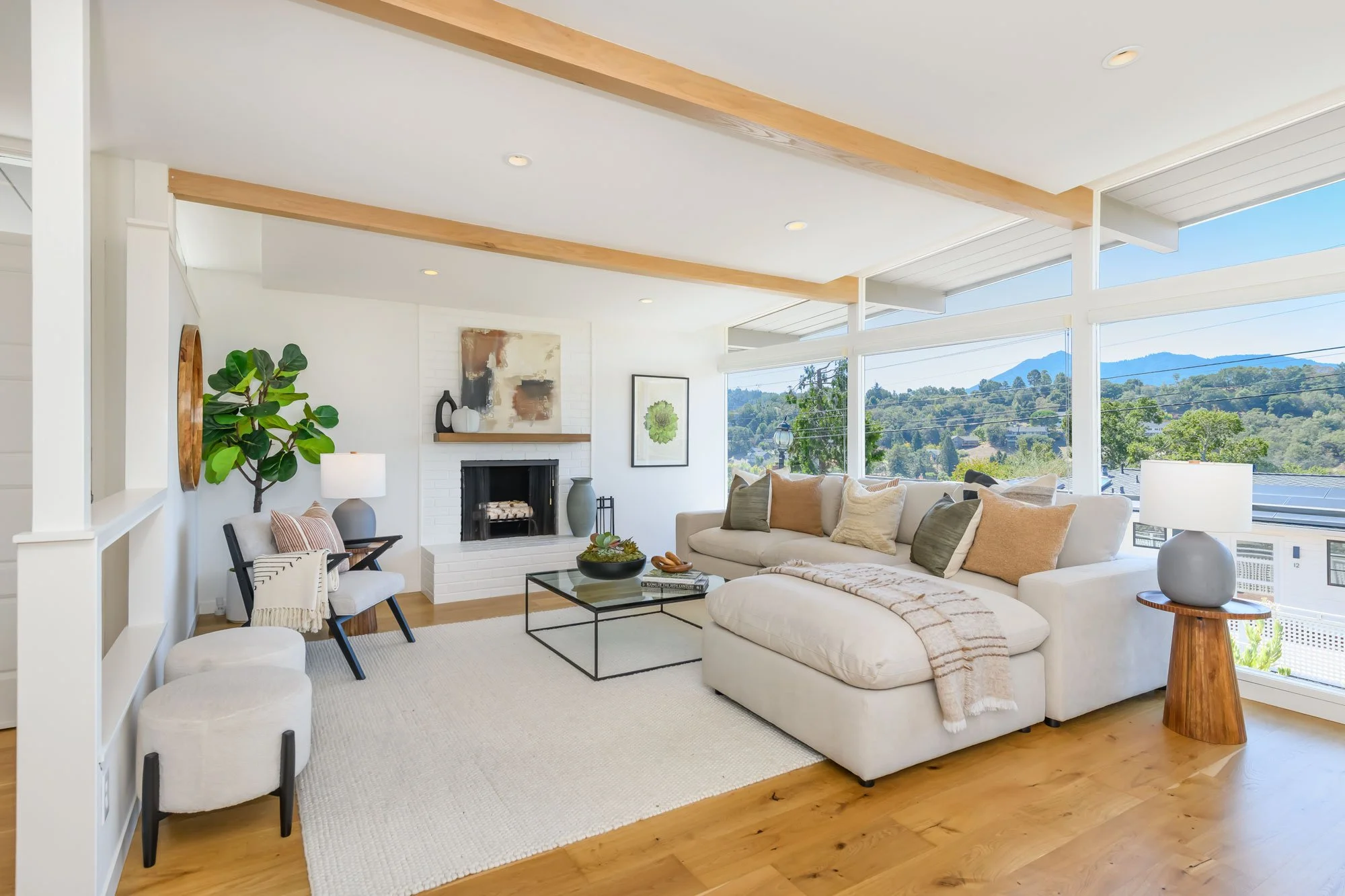 Bright living room with large windows showing a view of trees and mountains, beige sofa with multiple pillows, modern artwork above a fireplace, green plant, and contemporary furniture accents.