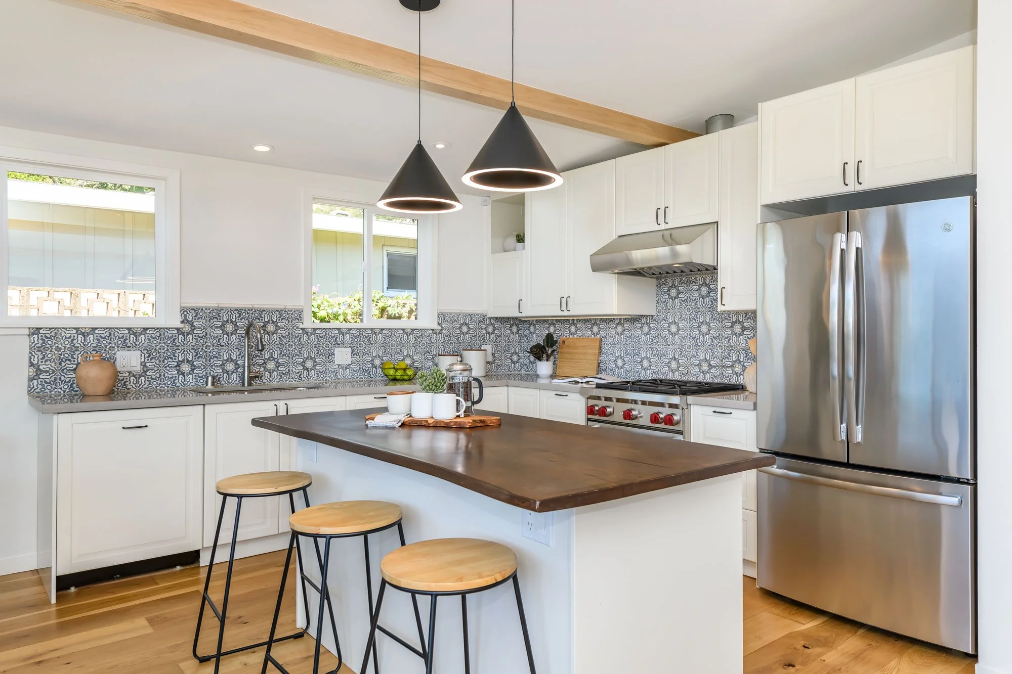 Modern kitchen with white cabinets, stainless steel refrigerator, patterned tile backsplash, wooden countertops, black pendant lights, and wooden bar stools around a kitchen island.