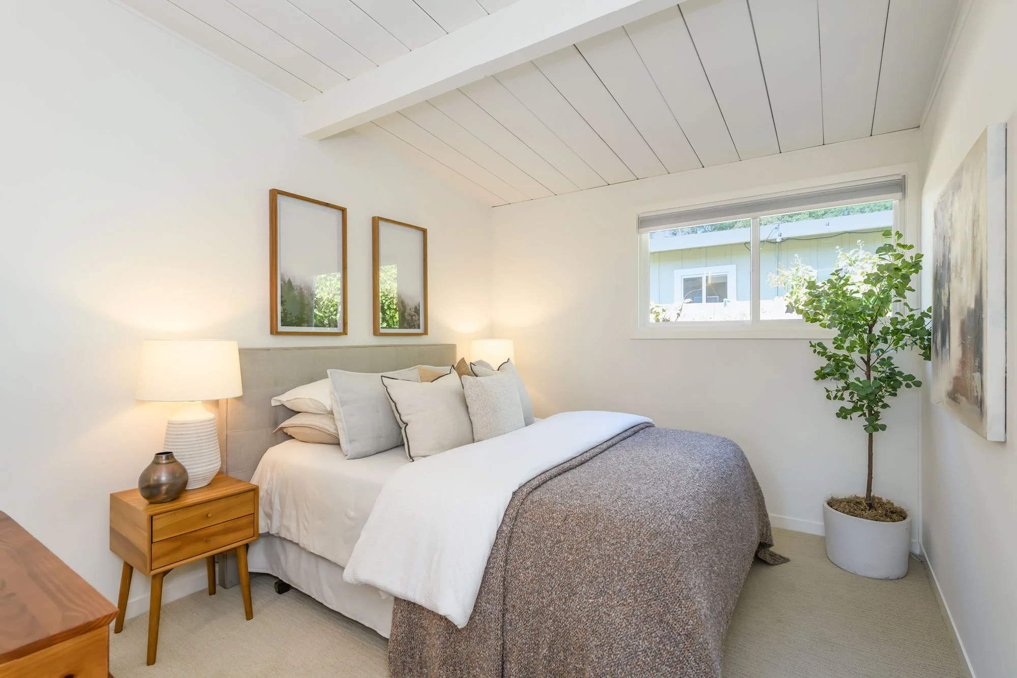 Bright bedroom with white walls, a bed with white and gray bedding, a small wooden nightstand with a lamp, framed artwork on the wall, a large window with blinds, and a potted plant in the corner.