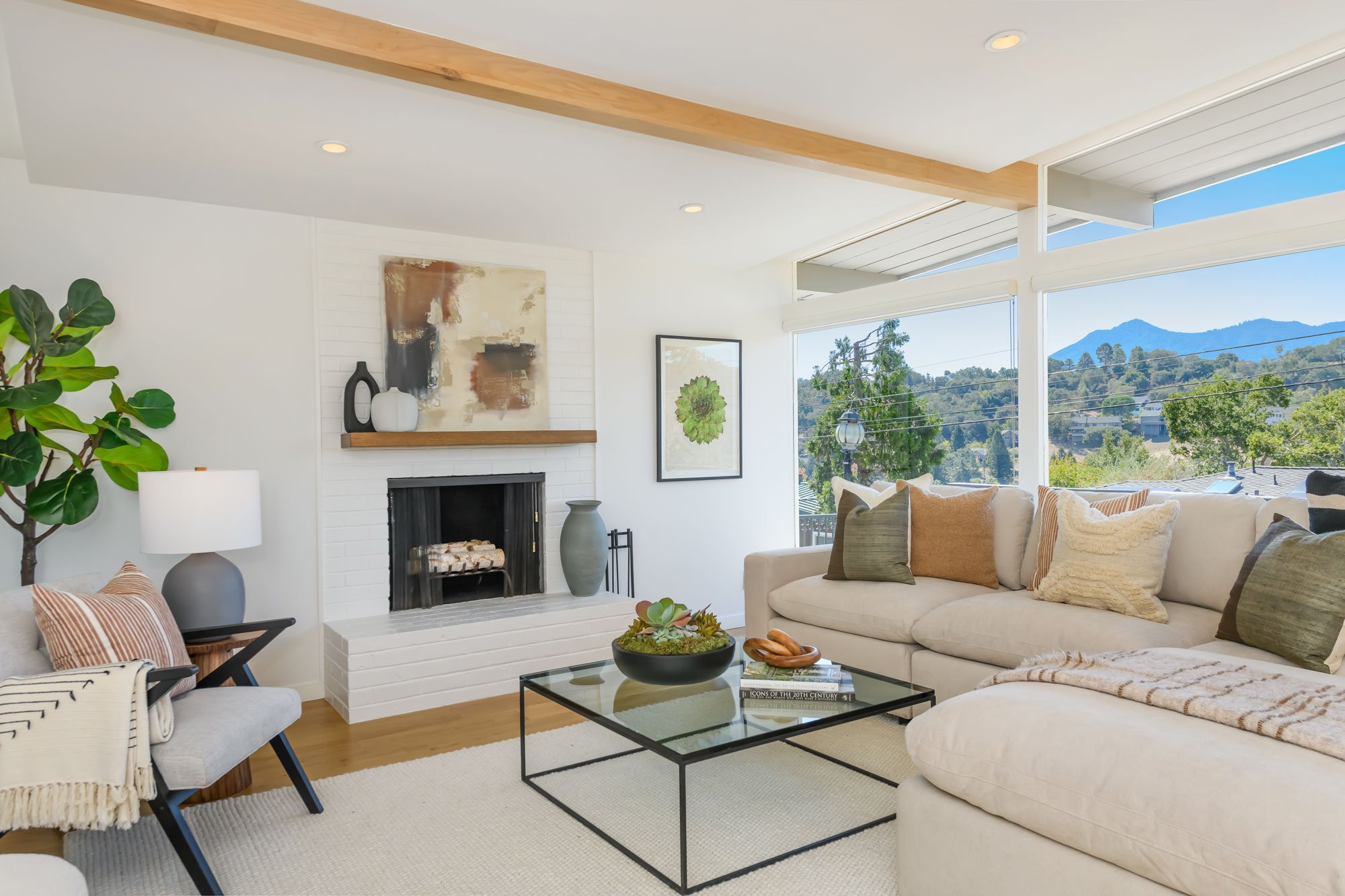Bright living room with white walls, large windows showing green trees and hills, beige sectional sofa with pillows, glass coffee table with books and a potted plant, fireplace with decorative vases, abstract artwork, and a large potted plant.