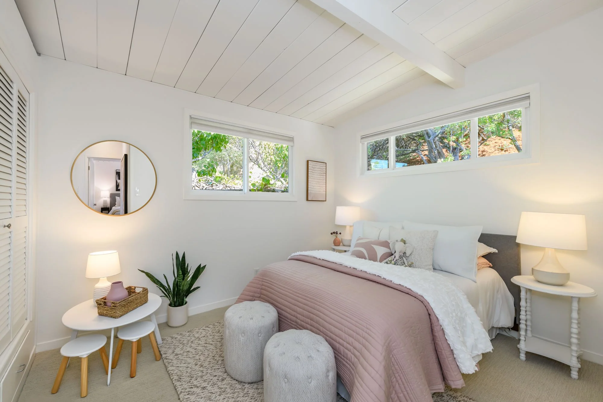 A cozy bedroom with white walls, a bed with pink and white bedding, two white ottomans, and bedside lamps. There is a small white table with a basket, purple vases, and a plant. The room has two large windows showing outside greenery, and a circular 