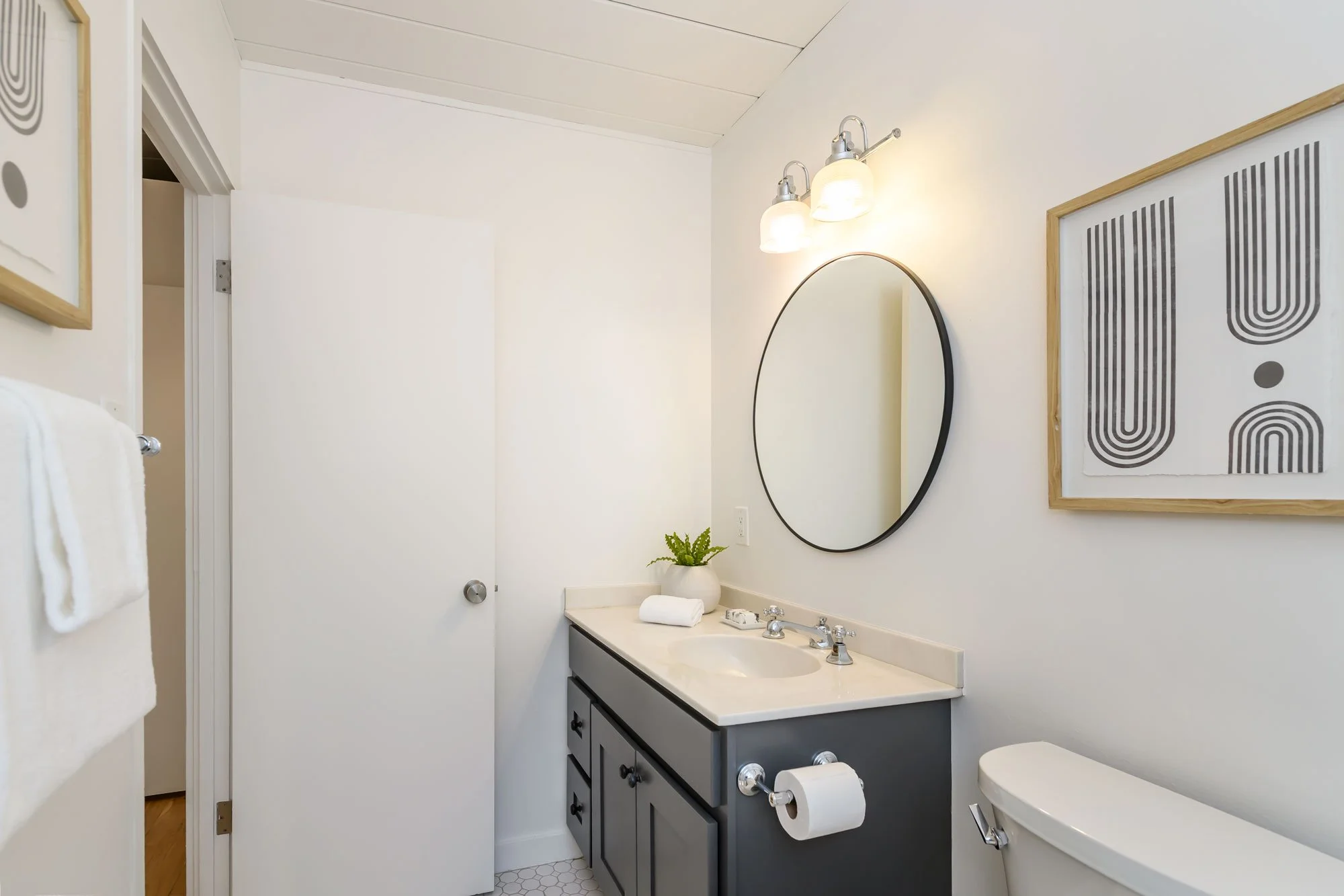 Contemporary bathroom with white walls, wooden framed black and white artwork, a large oval mirror, wall-mounted light fixtures, a black vanity with a white countertop, a small potted fern, neatly folded towels, and bathroom essentials.