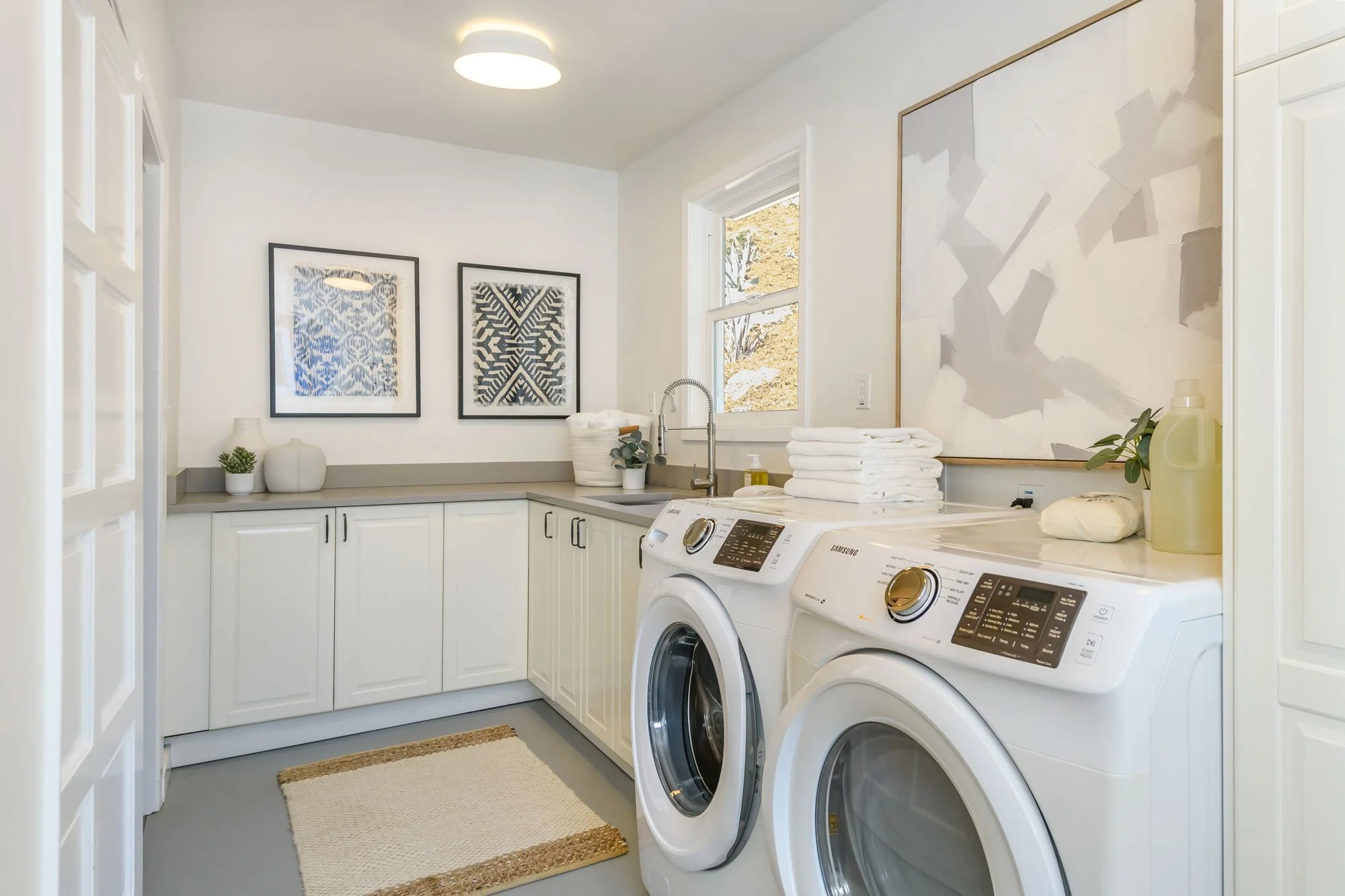 A laundry room with white cabinets, a window, wall art, and a stack of folded towels. There are two white washing machines. Decor includes small potted plants and bottles of laundry detergent.