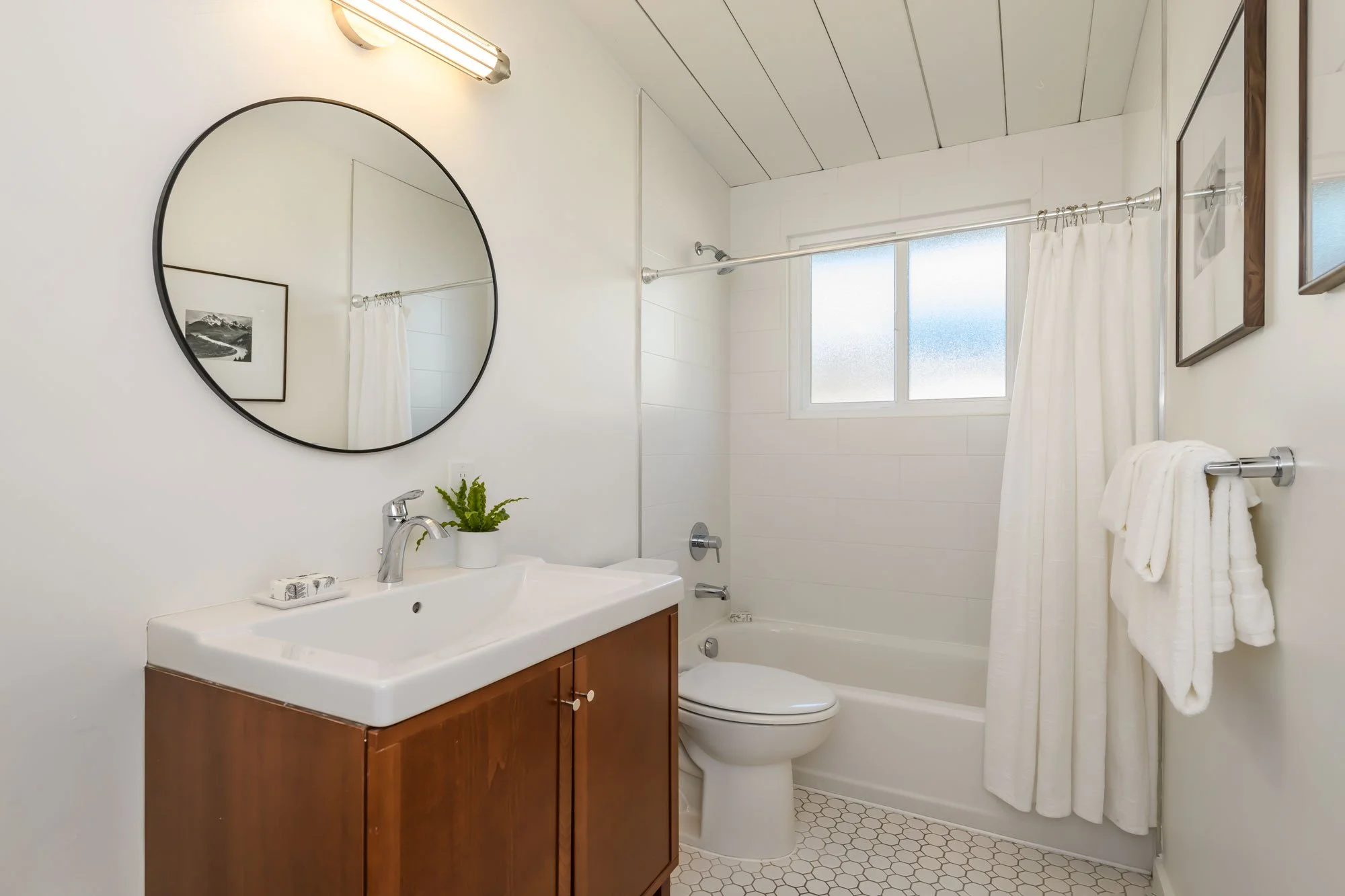 A small bathroom featuring a white sink with a wooden cabinet below, a round mirror above the sink, a toilet next to the sink, and a bathtub with a white shower curtain. There is a small potted plant on the sink, framed artwork on the wall, and a win