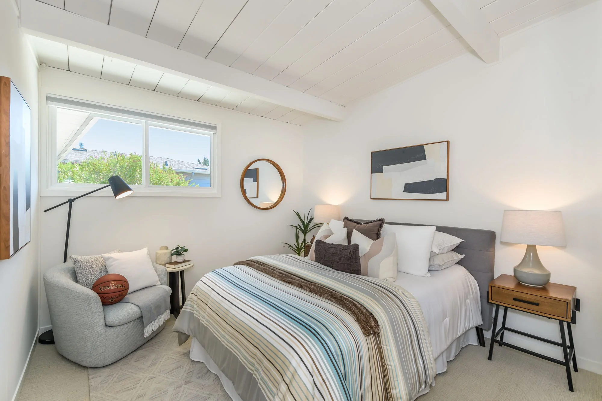 A bedroom with white walls, a sloped ceiling, and a large window showing trees outside. The bed has a striped comforter and multiple pillows. A bedside table with a lamp is next to the bed. There is a round mirror on the wall and abstract artwork abo