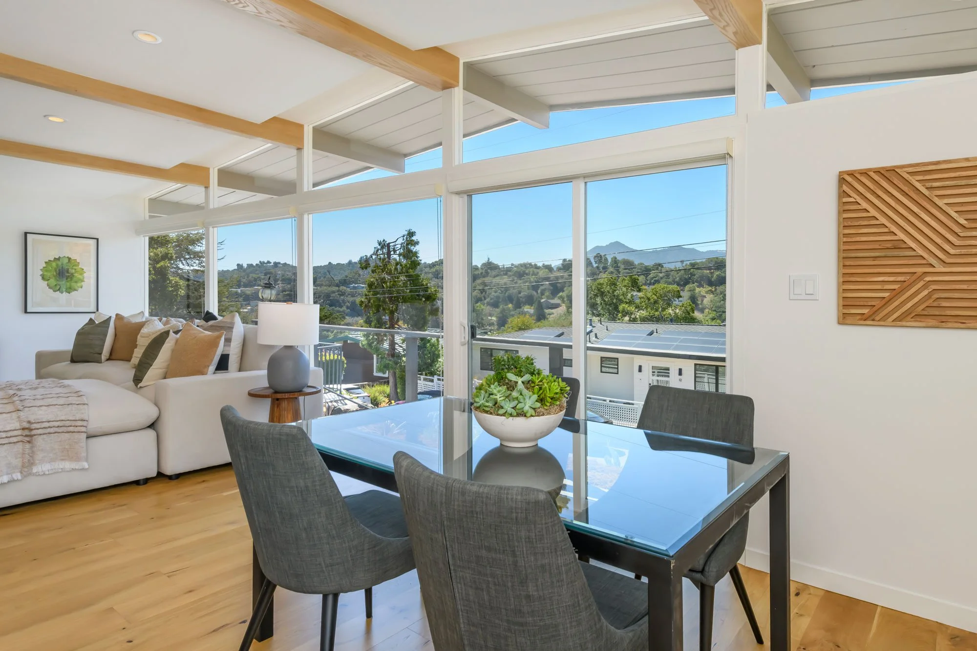 Living and dining room with large windows showing a scenic view of trees and mountains, a white sofa with multiple pillows, a glass dining table with four gray chairs, a decorative wooden wall panel, artwork on the wall, and plants on the table.