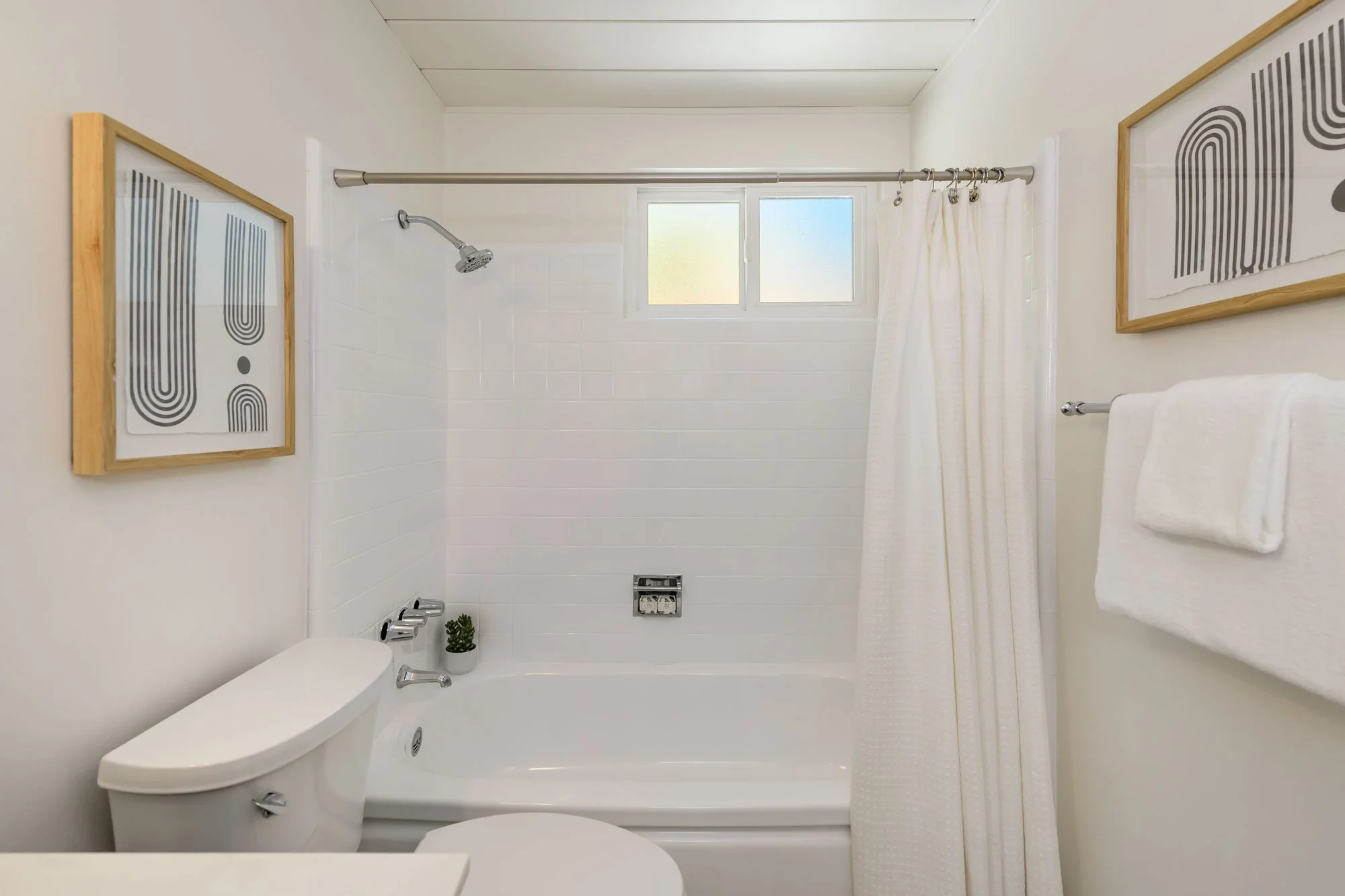 A modern bathroom with a white bathtub, toilet, and a small green plant. There are two framed abstract black and white art pieces on the wall, a white curtain on a rod, and a small frosted window above the bathtub.