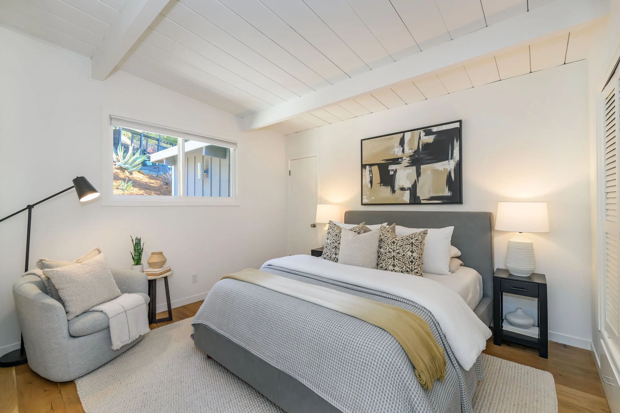A modern bedroom with a white ceiling and walls, a large window showing outdoor plants, a gray bed with decorative pillows, a white lamp on a black nightstand, a beige rug, and a small seating area with a white armchair, pillows, and a side table.