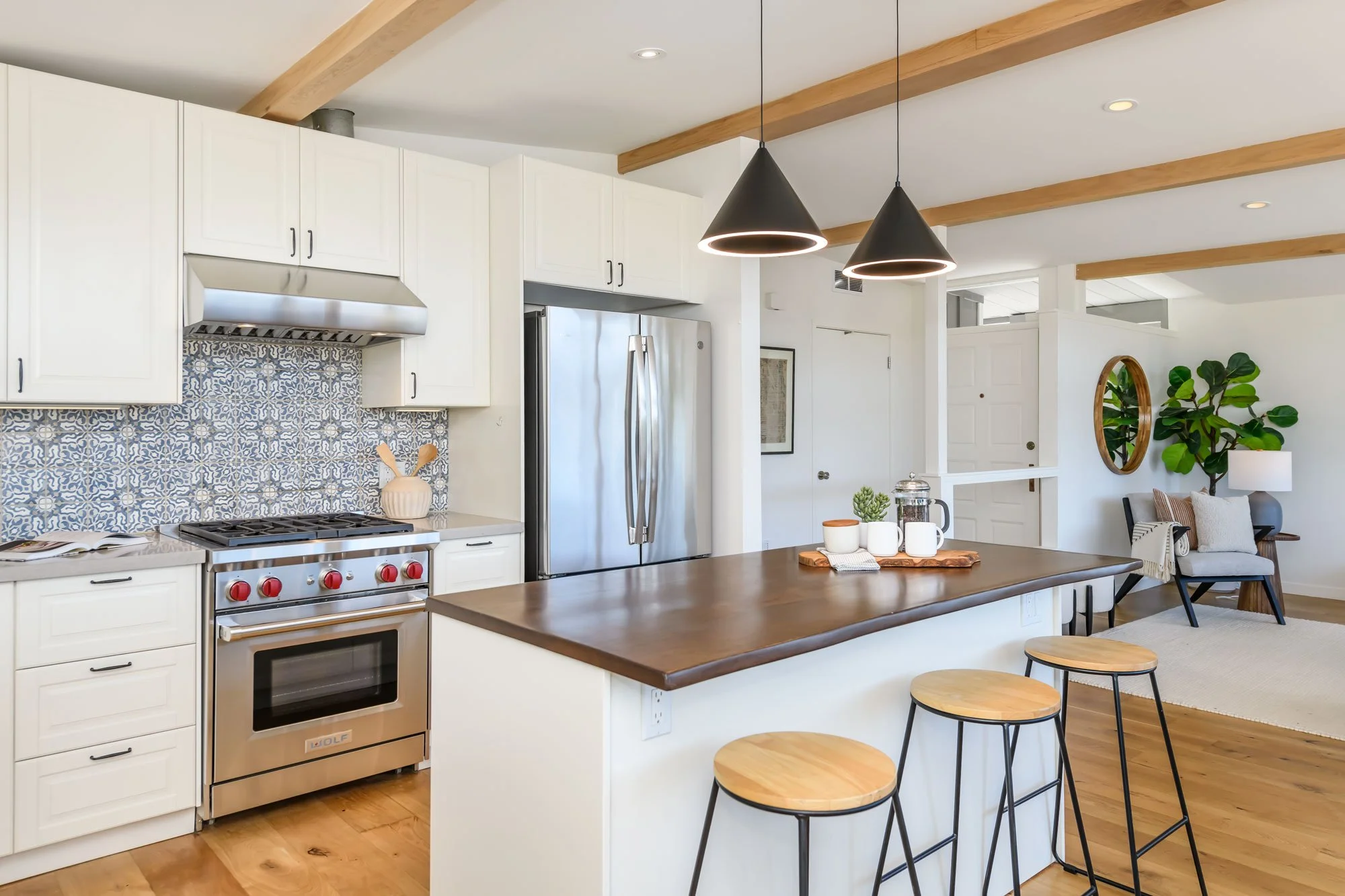 Modern kitchen with white cabinets, a stainless steel refrigerator, a patterned backsplash, a wooden dining table with cups, and black stools with wooden seats. Pendant lights hang from the ceiling, and a living room with a green plant, a mirror, and