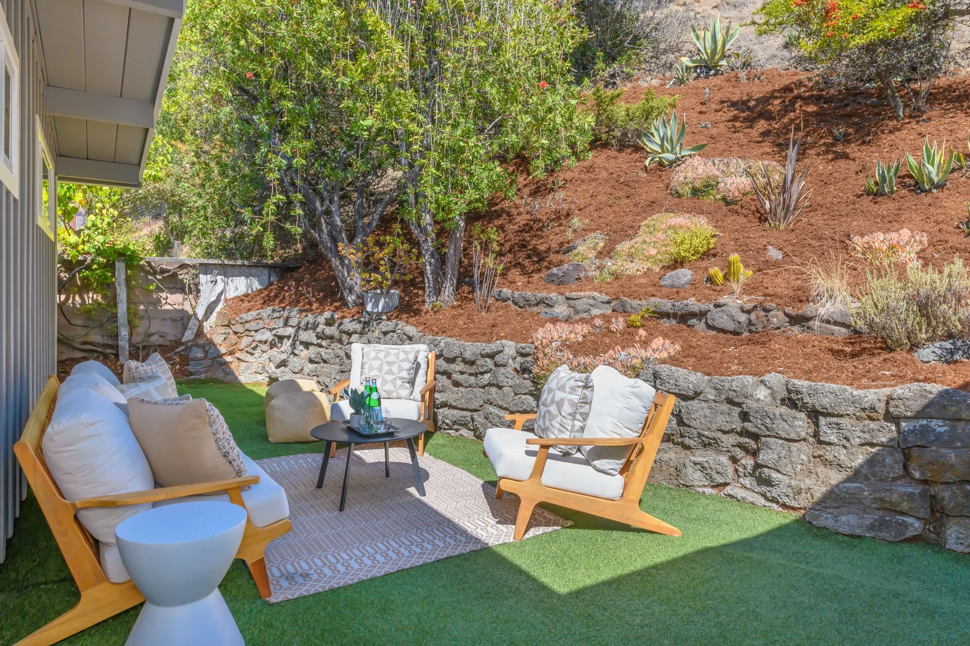 Outdoor patio with modern furniture, cushions, and small table on artificial grass, overlooking a landscaped garden with trees, shrubs, and rocks.