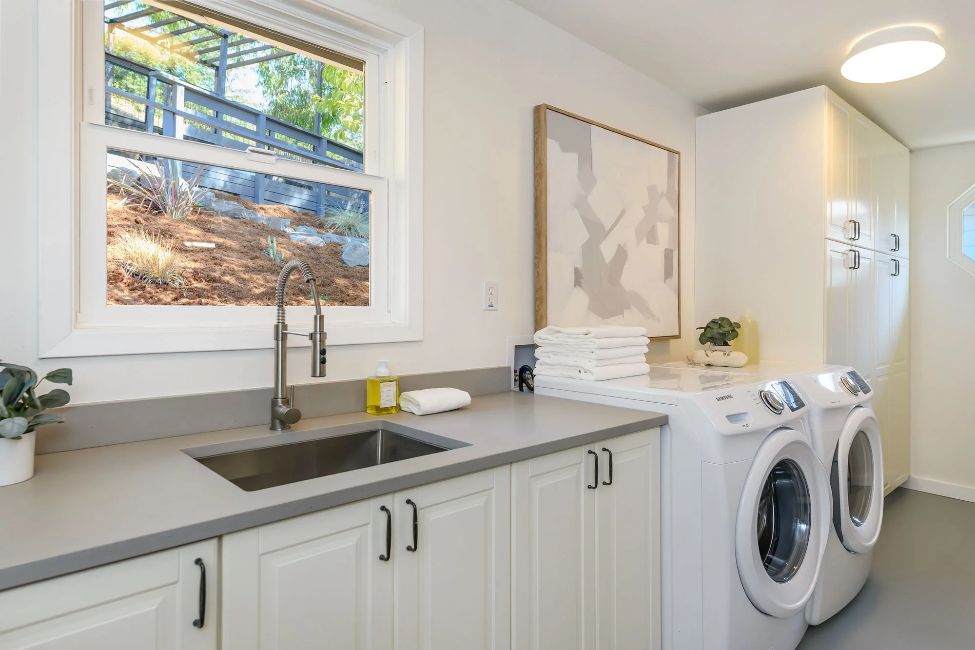 A laundry room with a sink, stack of folded towels, a washing machine, a dryer, and a window showing an outdoor landscaped area with plants and a fence.