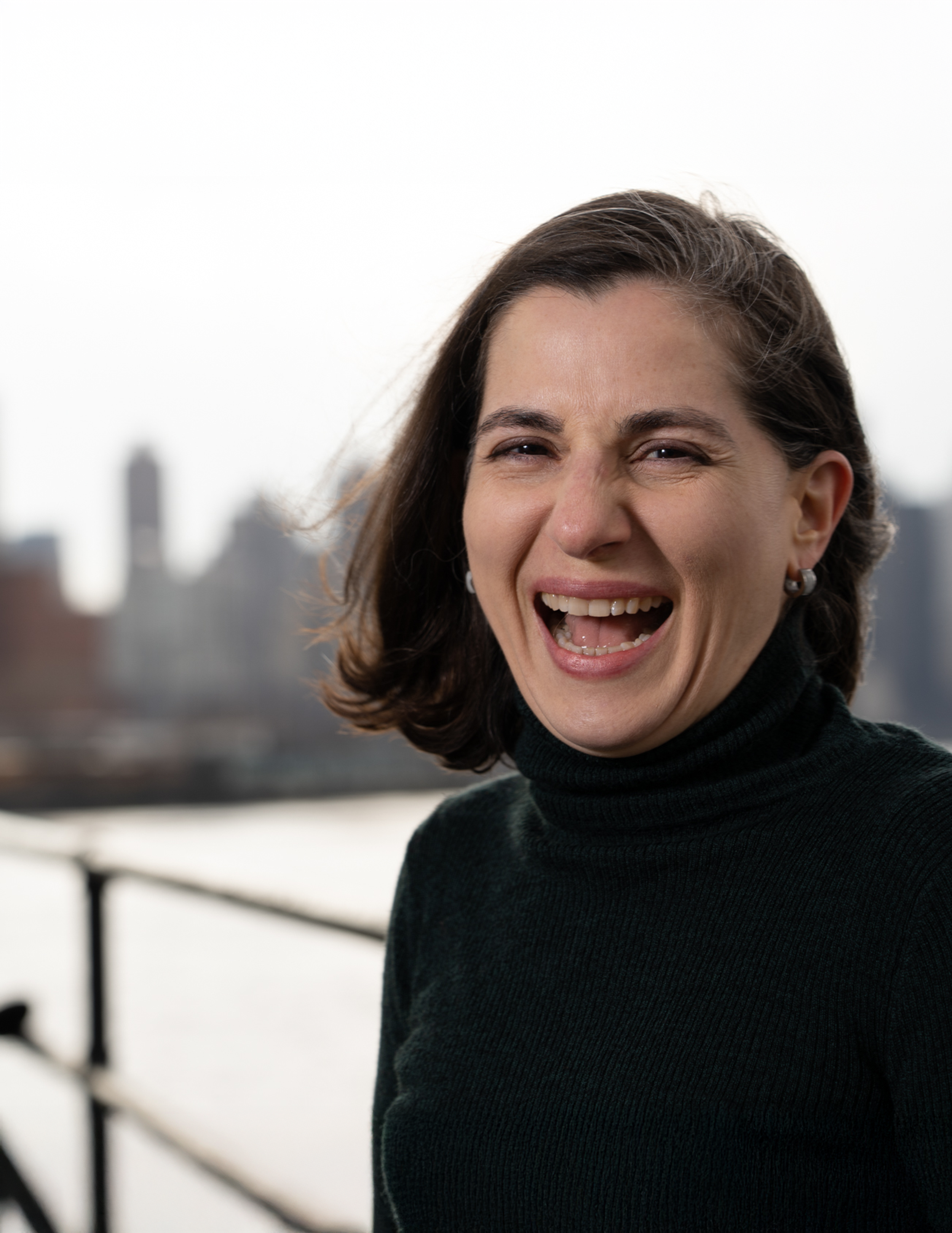 A woman with dark brown hair smiling and showing her teeth, wearing a black turtleneck, outdoors with an urban cityscape in the background.