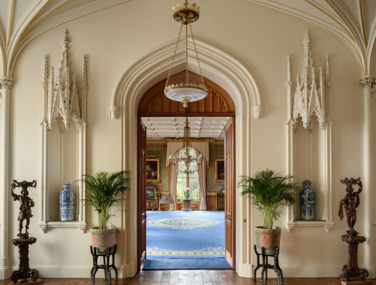 A grand hallway with ornate white molding, two potted plants, and decorative statues, leading into a room with large windows, chandeliers, and classical paintings.