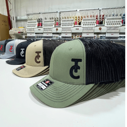 A row of baseball caps with different colors and a stitched logo, displayed on a table in front of embroidery machines.