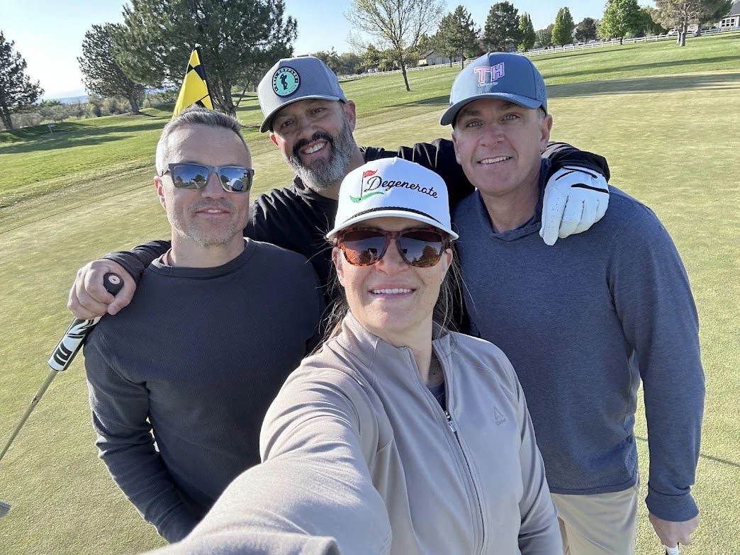 Four people taking a selfie on a golf course, smiling, with a flag and trees in the background. They are wearing casual golf attire and sunglasses.