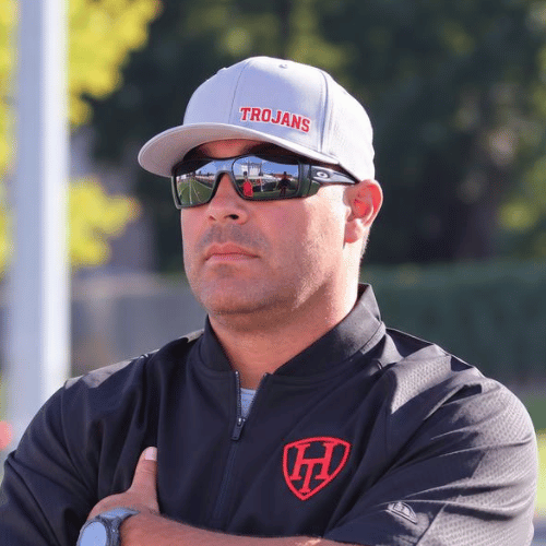 A man wearing a gray baseball cap with 'TROJANS' in red, sunglasses, a black jacket with a red shield logo, and a watch on his left wrist, standing outdoors with trees and a fence in the background.