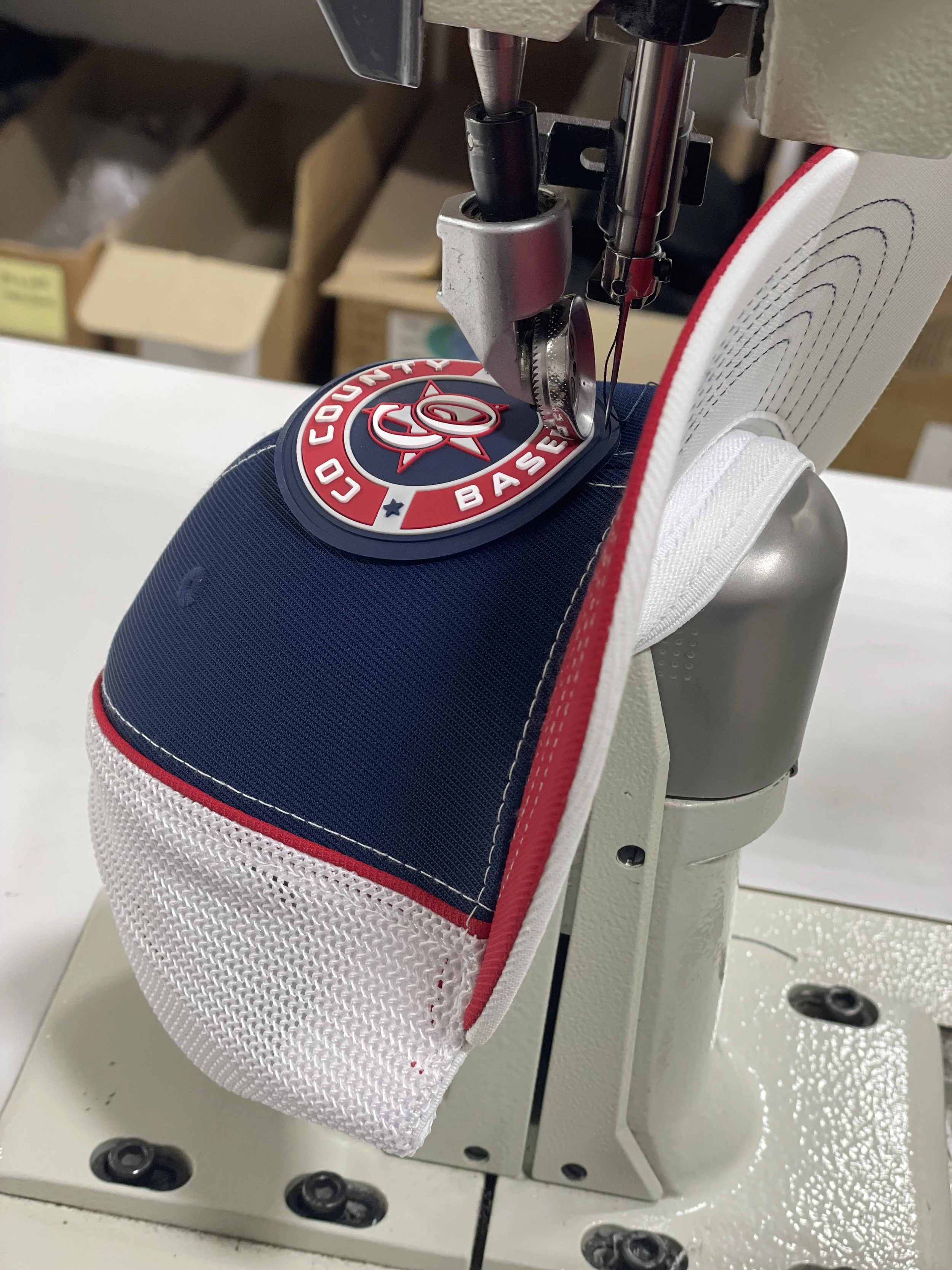 Close-up of a sewing machine process creating a patch being sewn on to a hat. The patch is blue with white mesh backing and is being sewn onto a white fabric with red and white trim.