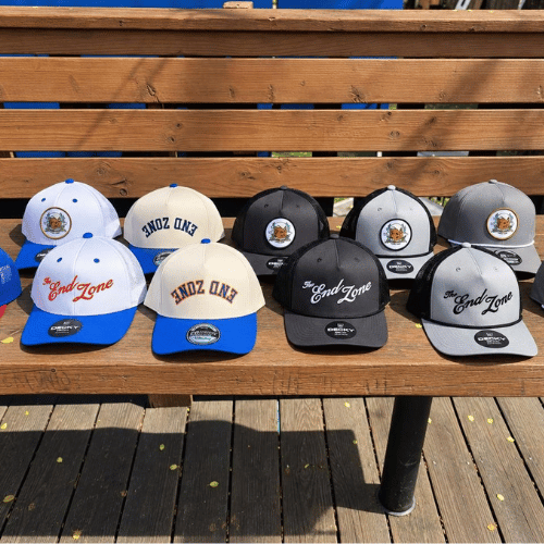 Six baseball caps on a wooden bench with a wooden fence in the background. The caps are in pairs of white and blue, black, and gray, with logo patches and embroidered text. Two caps have 