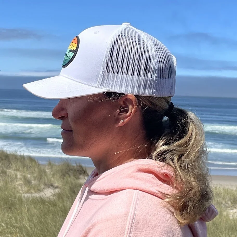 Woman in pink hoodie wearing a white cap with a colorful rainbow emblem, standing on a beach with sand dunes and the ocean in the background.