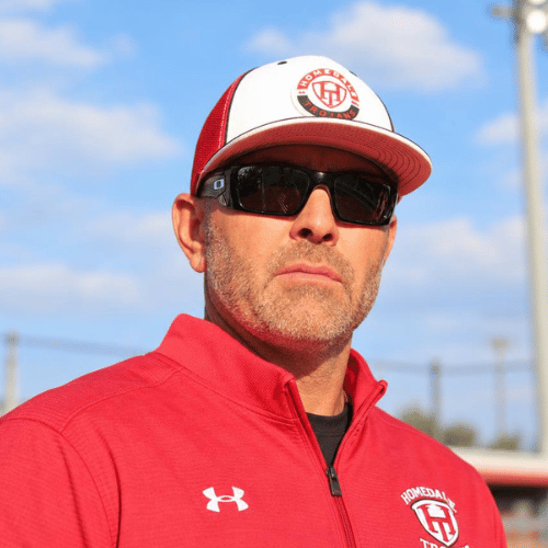 Man wearing a red jacket and a white and red baseball cap with a logo, sunglasses, outdoors with a blue sky and some utility poles in the background.