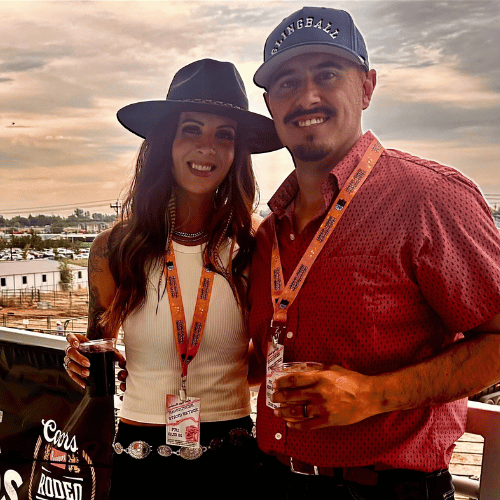A man and woman standing outdoors at sunset, both smiling and holding drinks. The woman wears a wide-brimmed hat and white top, while the man wears a baseball cap and red shirt. They have event passes around their necks.