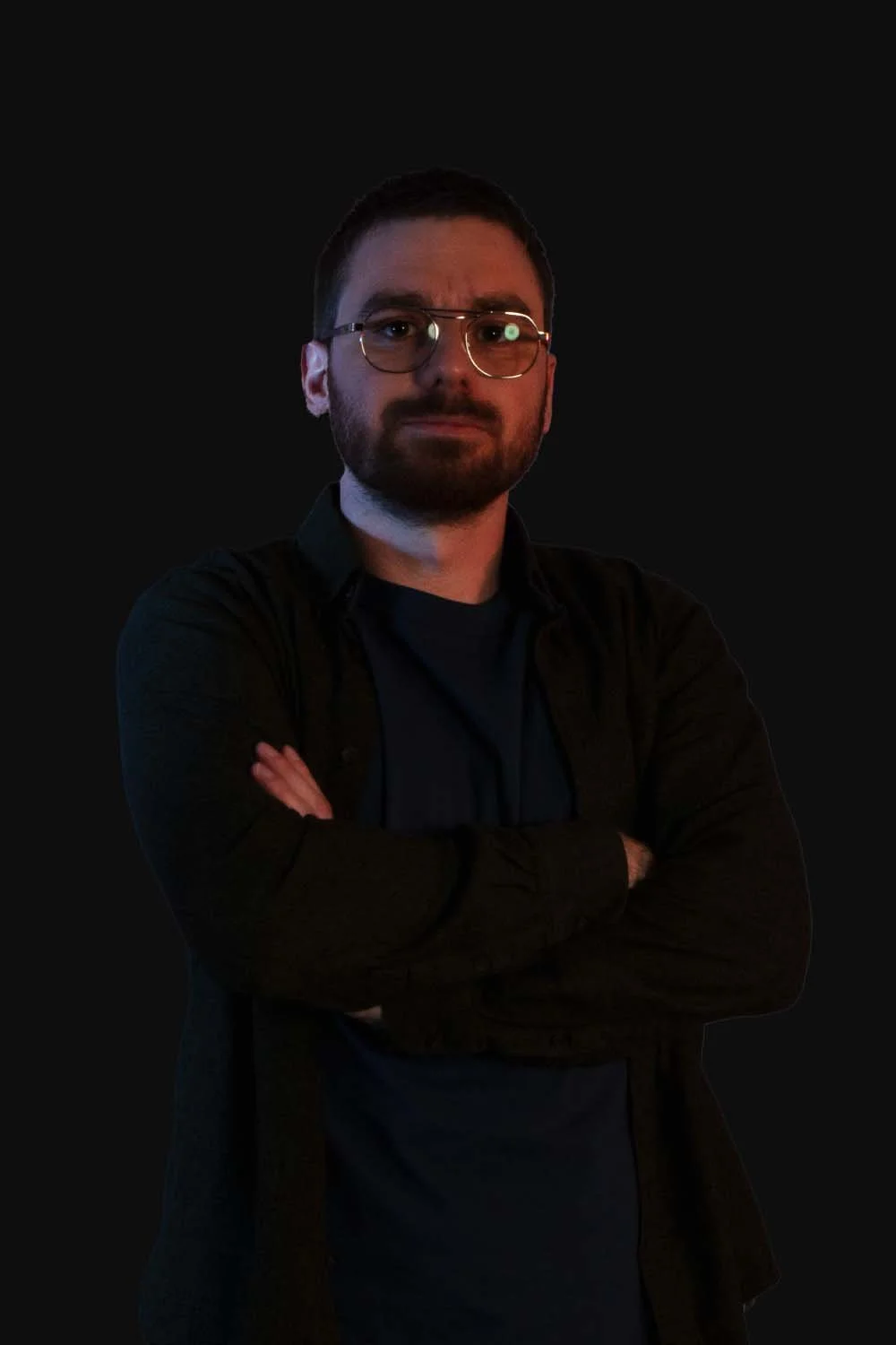 A man with glasses and a beard standing with arms crossed against a black background.