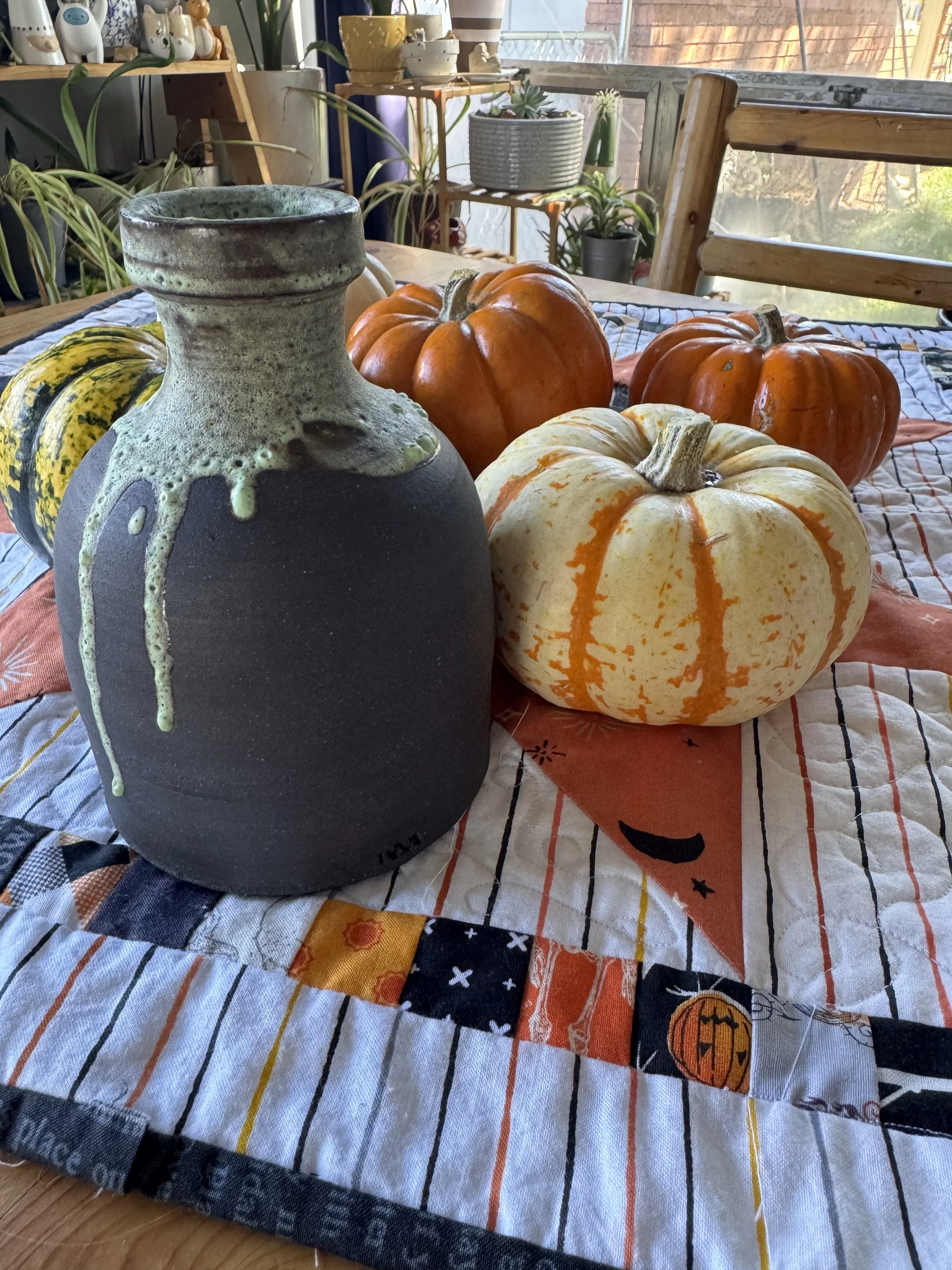 Various pumpkins and a ceramic vase on a table with a Halloween-themed cloth, inside a room with potted plants and shelves.