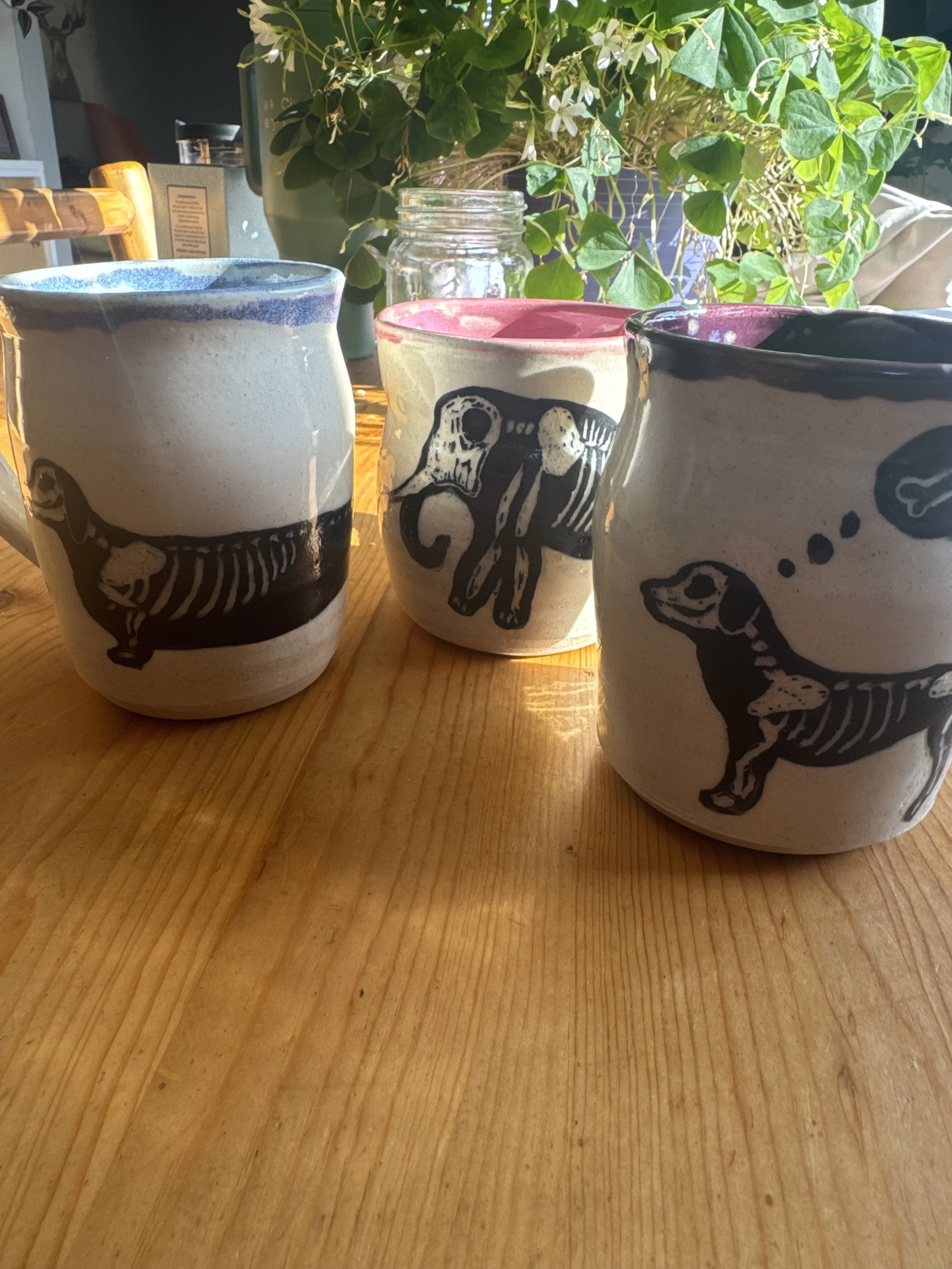 Three ceramic mugs with black skeletal animal designs, placed on a wooden table, with green plants and a glass jar in the background.