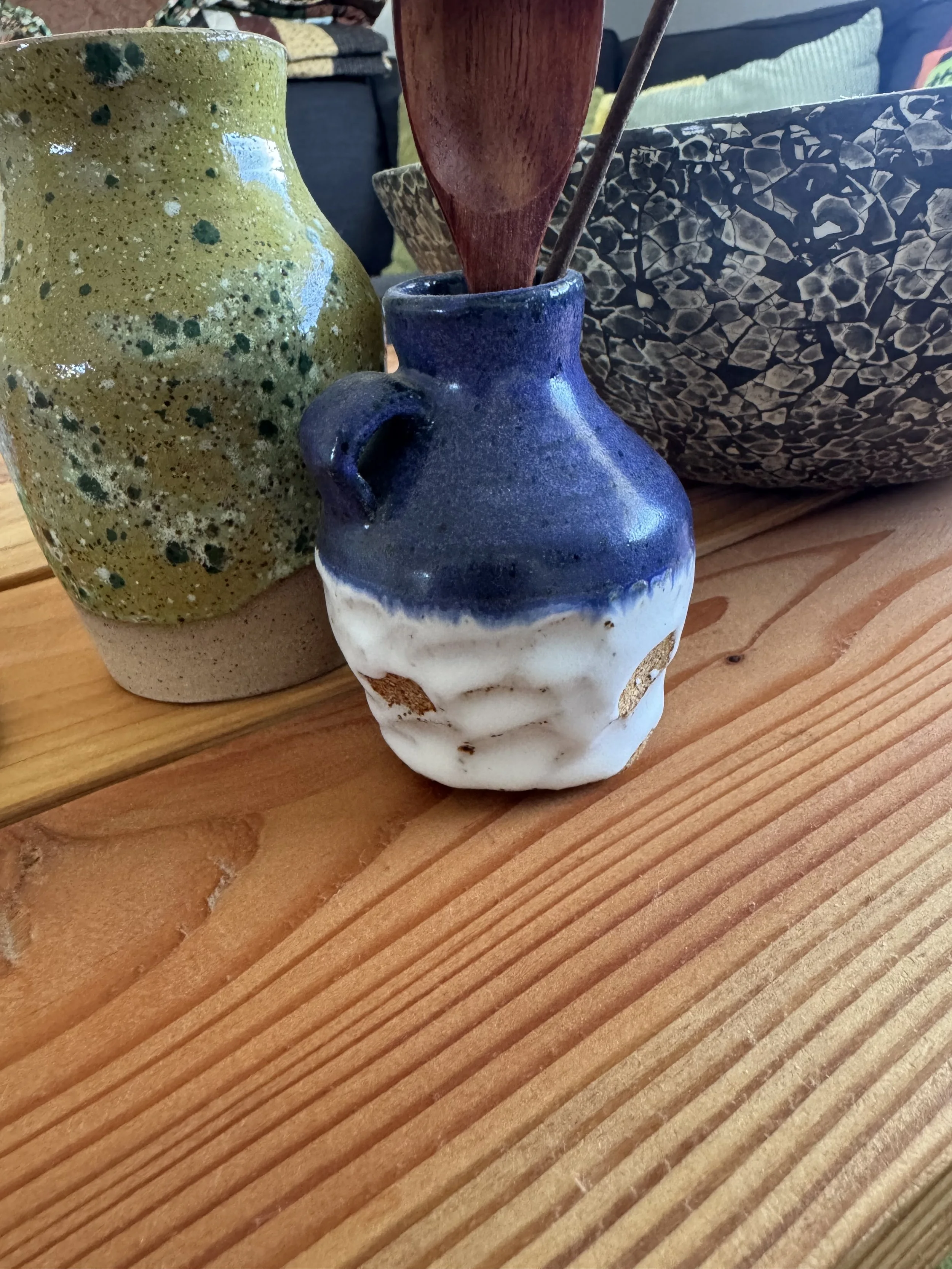 Close-up of three ceramic vases and a decorative bowl on a wooden surface. The left vase is green with darker green splatters, the middle is dark blue with white at the bottom, and the right bowl has a black and beige pattern.