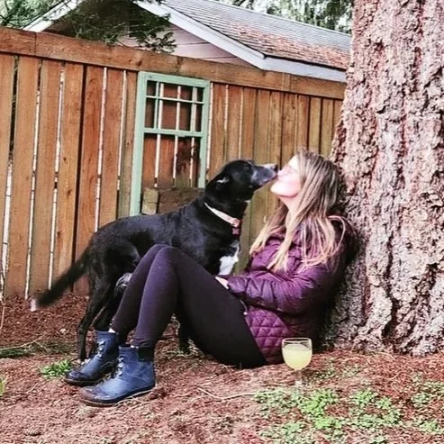 A woman sitting against a tree with a black dog giving her a kiss, in a backyard with a wooden fence and a small windowed structure.