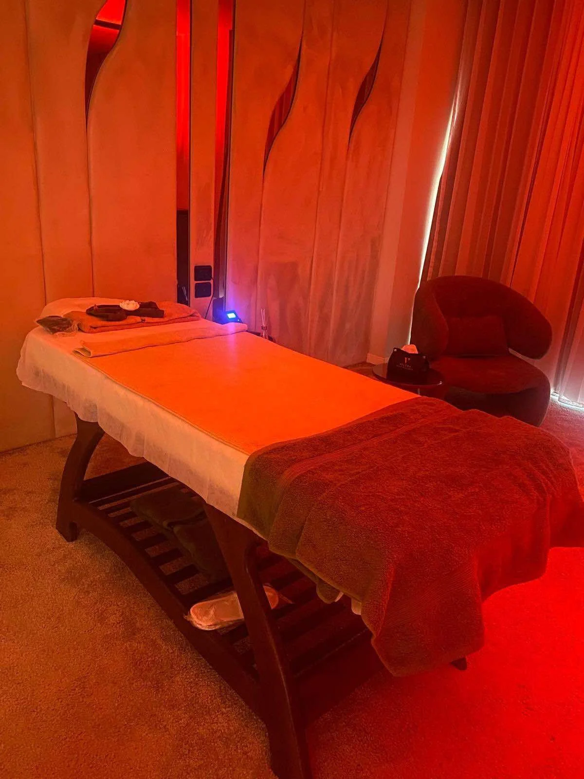 Treatment room with massage table covered in orange sheet, a towel with toiletries on top, a black chair, and red ambient lighting.
