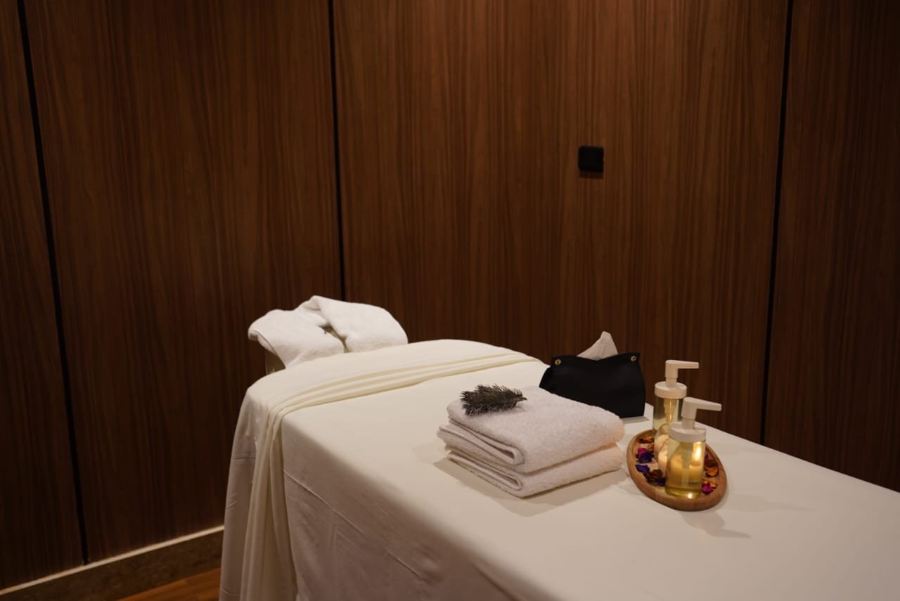 Spa massage table with folded towels, lotion bottles, and a black pouch, set against a wooden wall background.