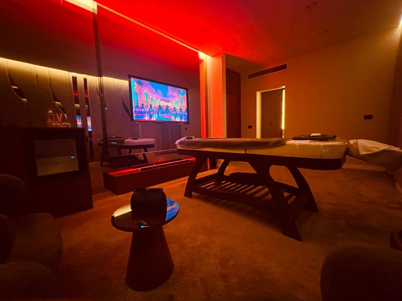 Dimly lit room with a lying massage table, a television displaying colorful animated scene, and a small round table with a black object on top.