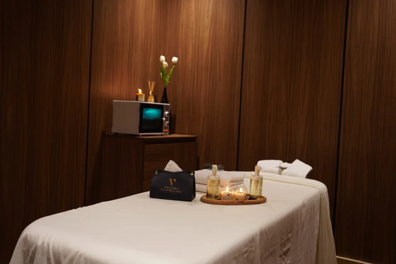 Spa or wellness treatment room with a massage table covered in a white sheet, surrounded by candles, lotions, and towels. The background features a wood-paneled wall, a small television, a vase with white flowers, and a black bag with the Velora Spa & Wellness logo.