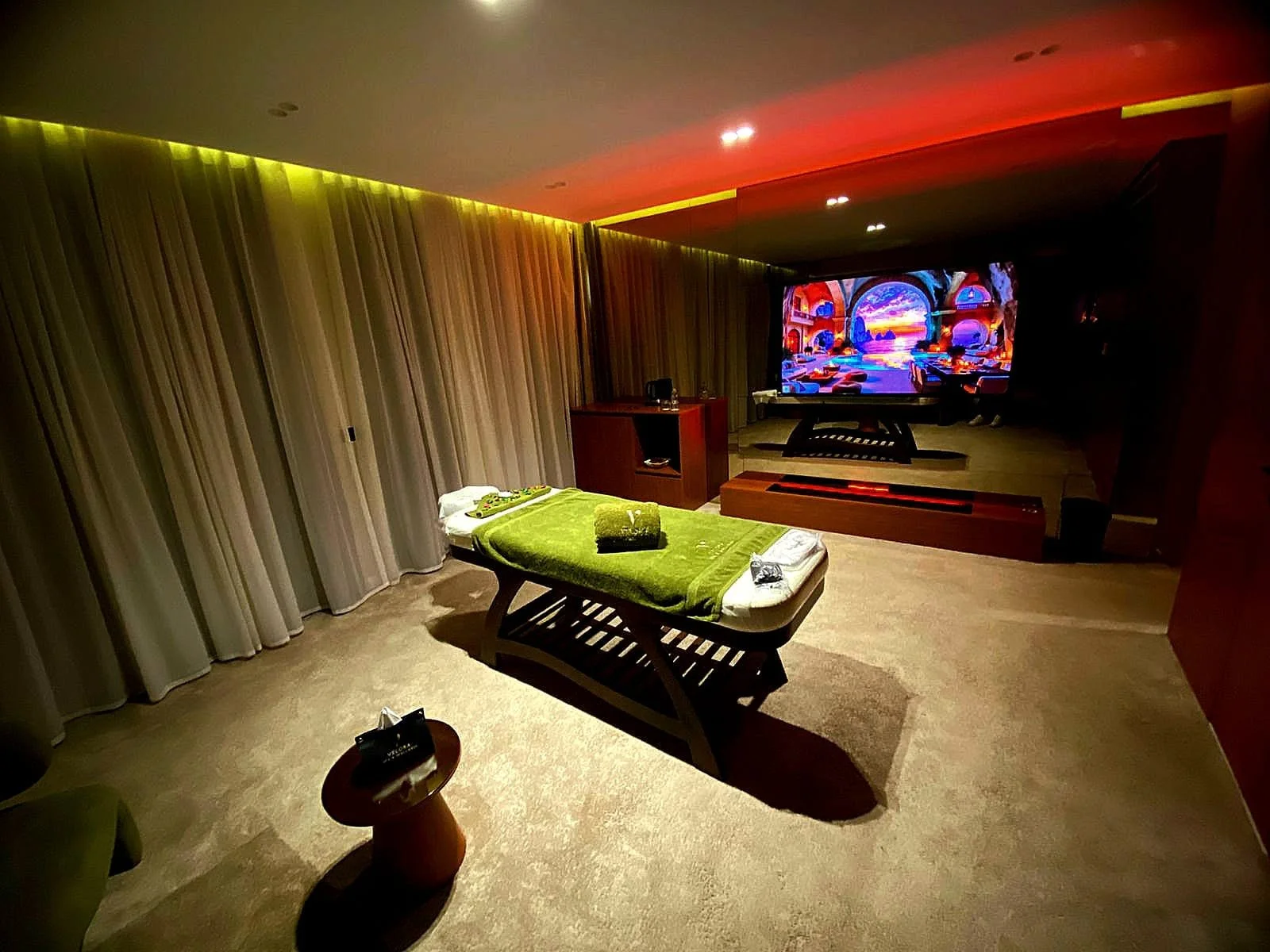 Hotel room with a single bed covered with green towels, a large flat-screen TV displaying colorful artwork, and neutral-colored curtains.