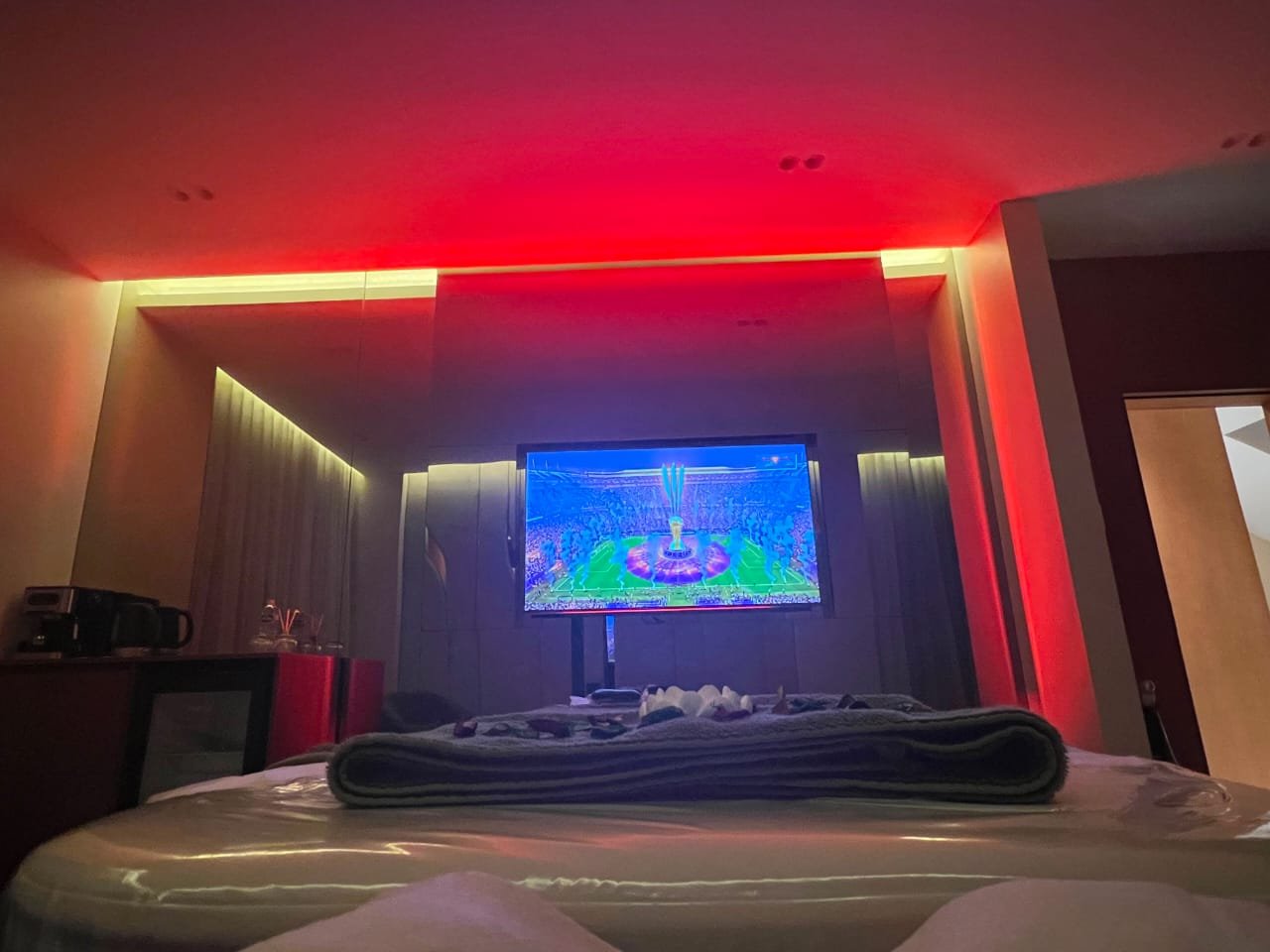 Hotel room with dim lighting, colorful LED accent lights on ceiling corners, a flat-screen TV displaying a sports game, and a bed with folded towels in the foreground.