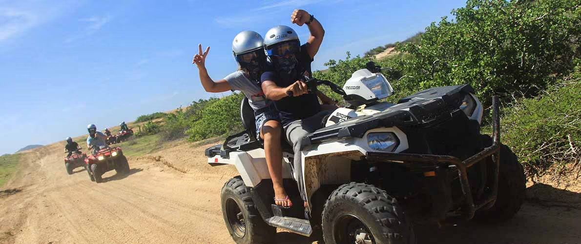 Two people riding an ATV on a dirt trail, wearing helmets and making peace signs, with additional ATV riders in the background and green bushes on the sides.