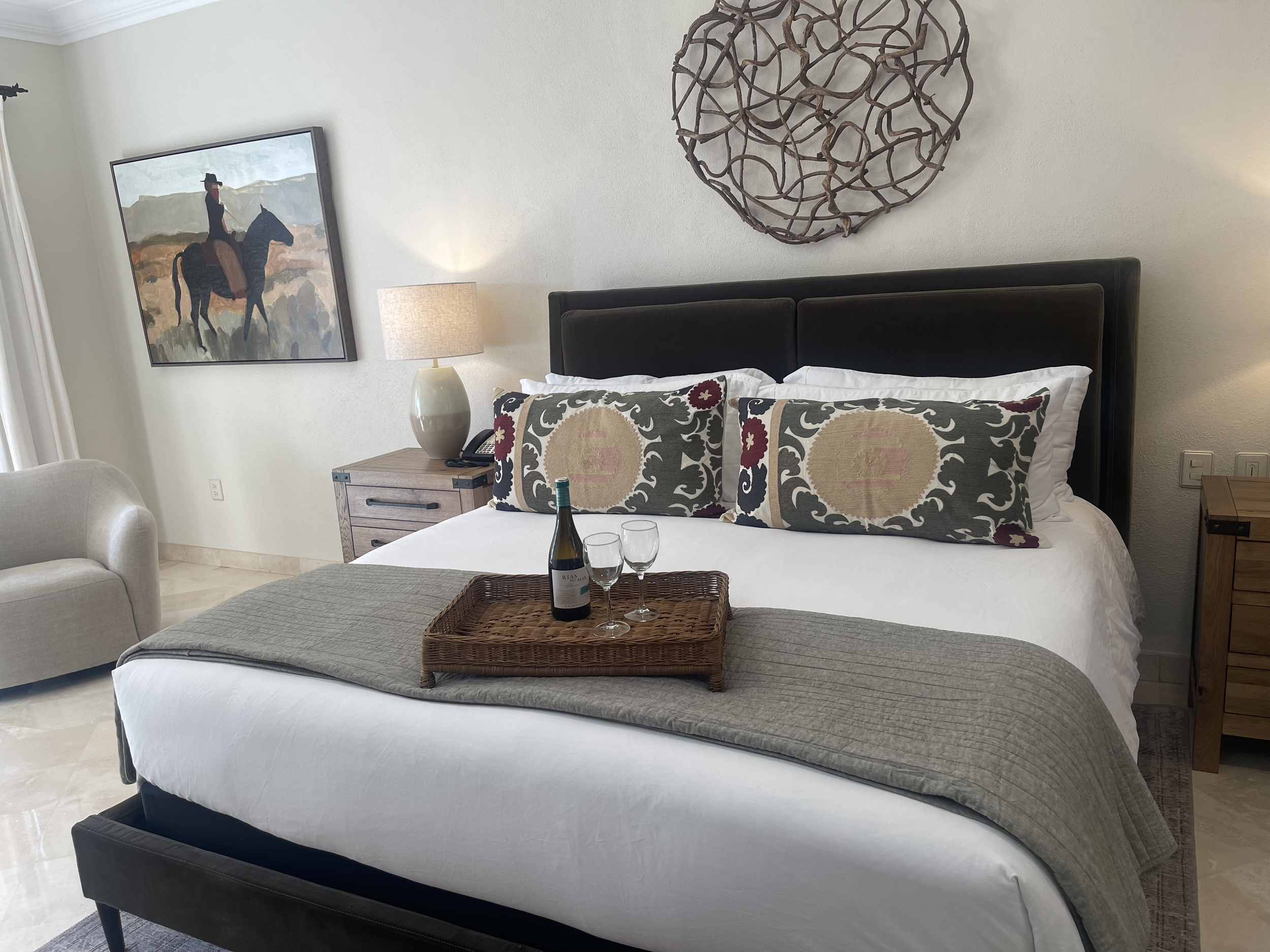 Condo room with a large bed decorated with vibrant red and patterned pillows, a nightstand with a lamp, a framed artwork on the wall, a sliding glass door leading to a balcony with chairs, and a view of palm trees outside.