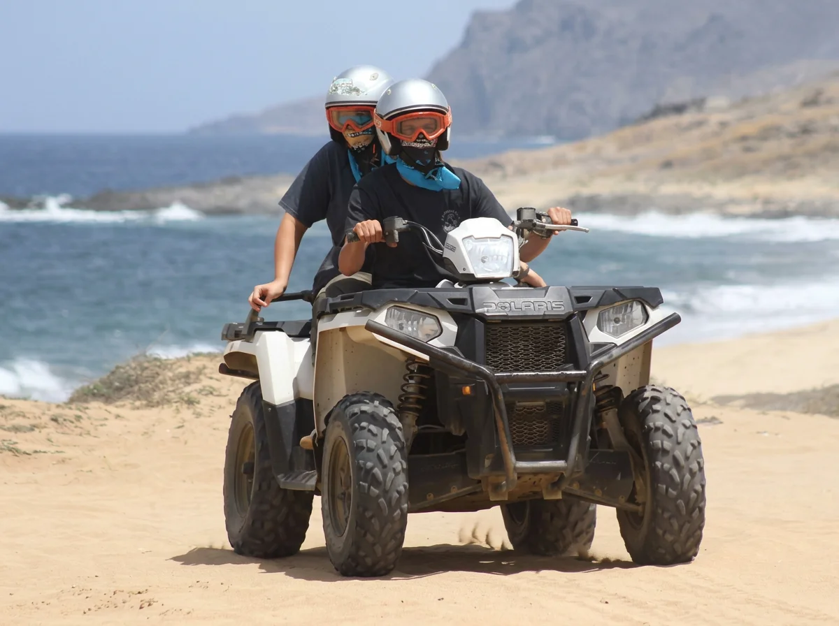 Two children riding an all-terrain vehicle (ATV) on a sandy beach with ocean and rocky cliffs in the background, both wearing helmets and face coverings.