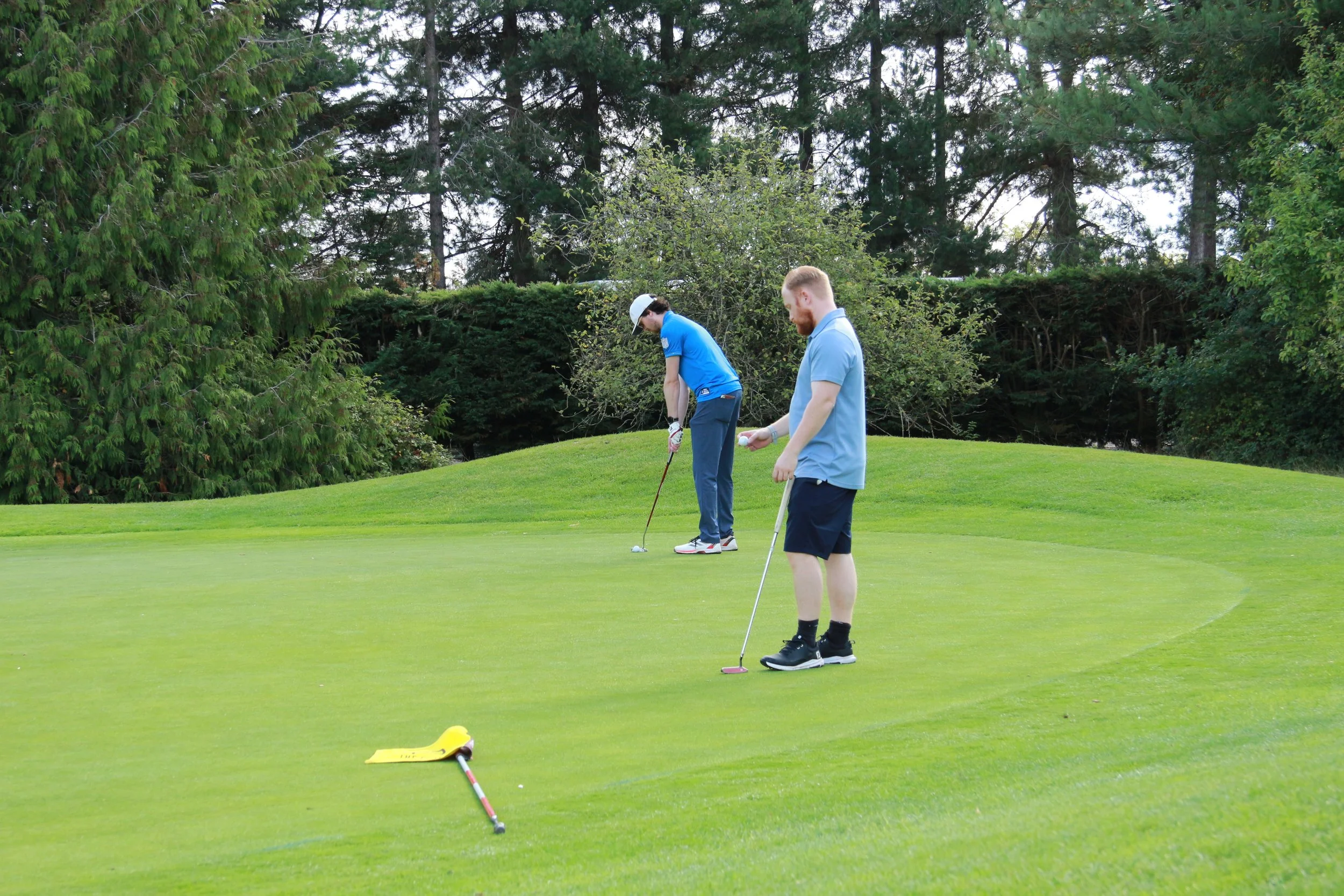 Two young men playing golf on a lush green course with tall trees in the background, one preparing to putt and the other holding a golf club.