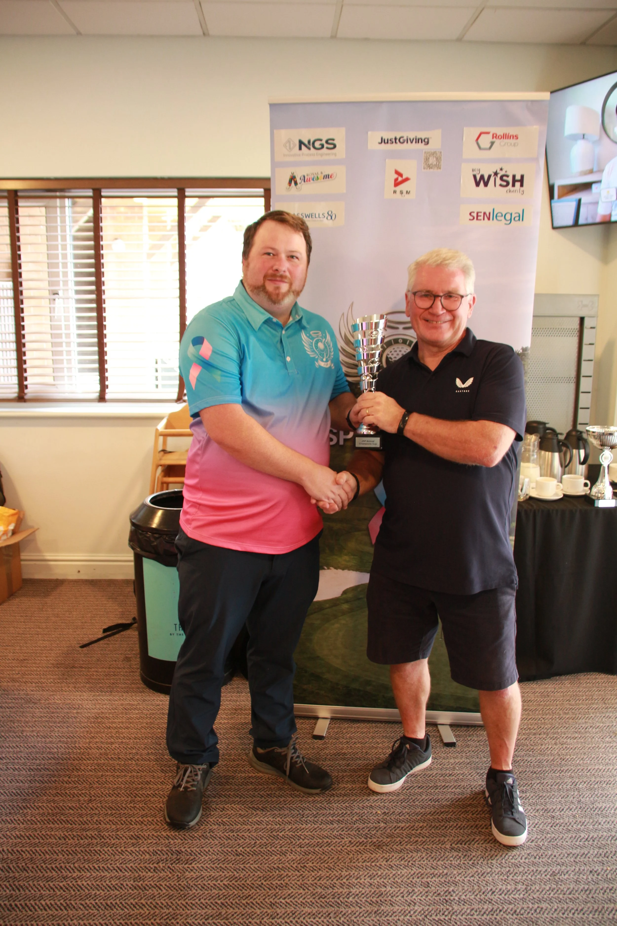 Two men shaking hands, one holding a trophy, standing in a room with a backdrop featuring various sponsor logos.