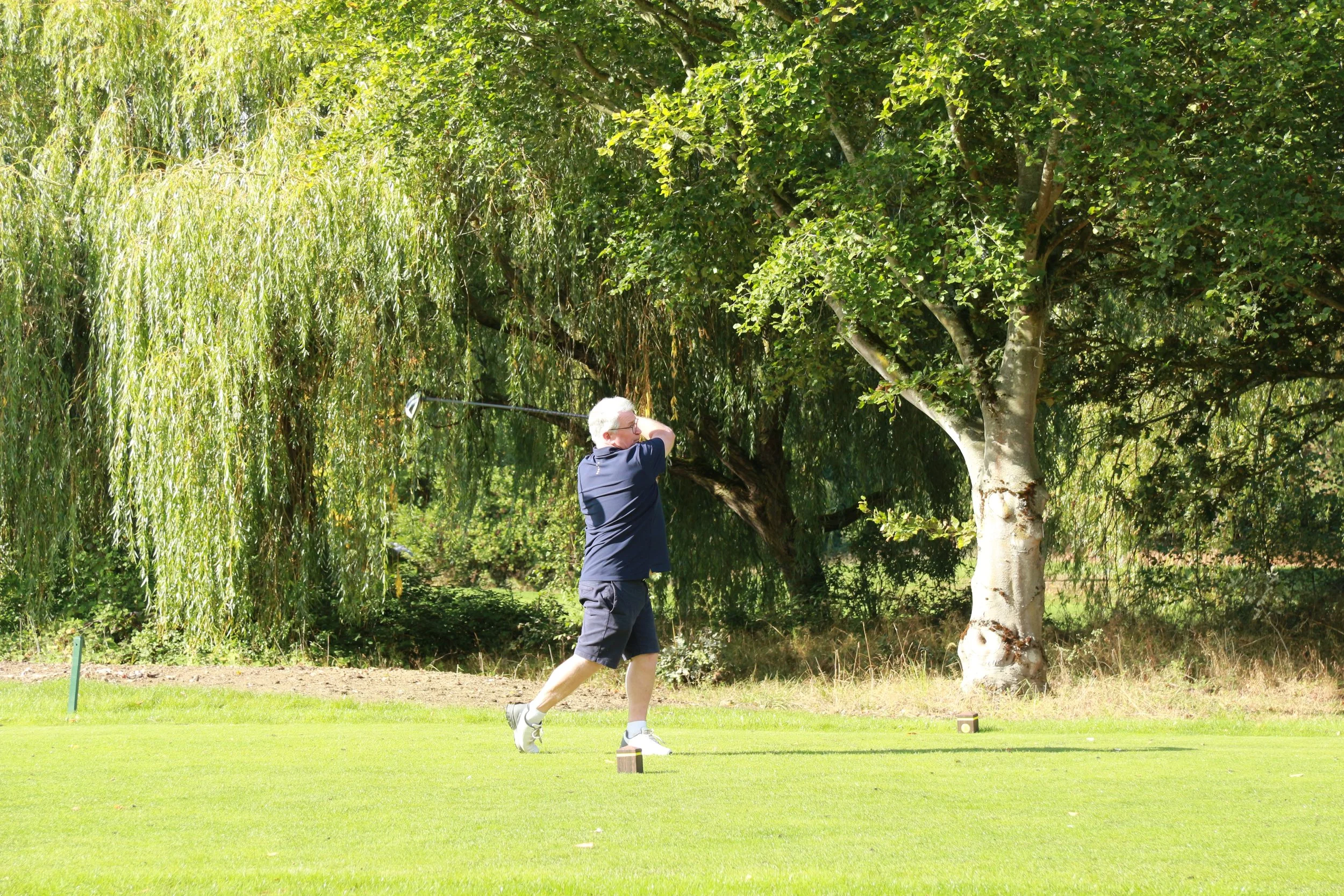 A man playing golf on a lush green course, swinging a golf club with trees in the background.