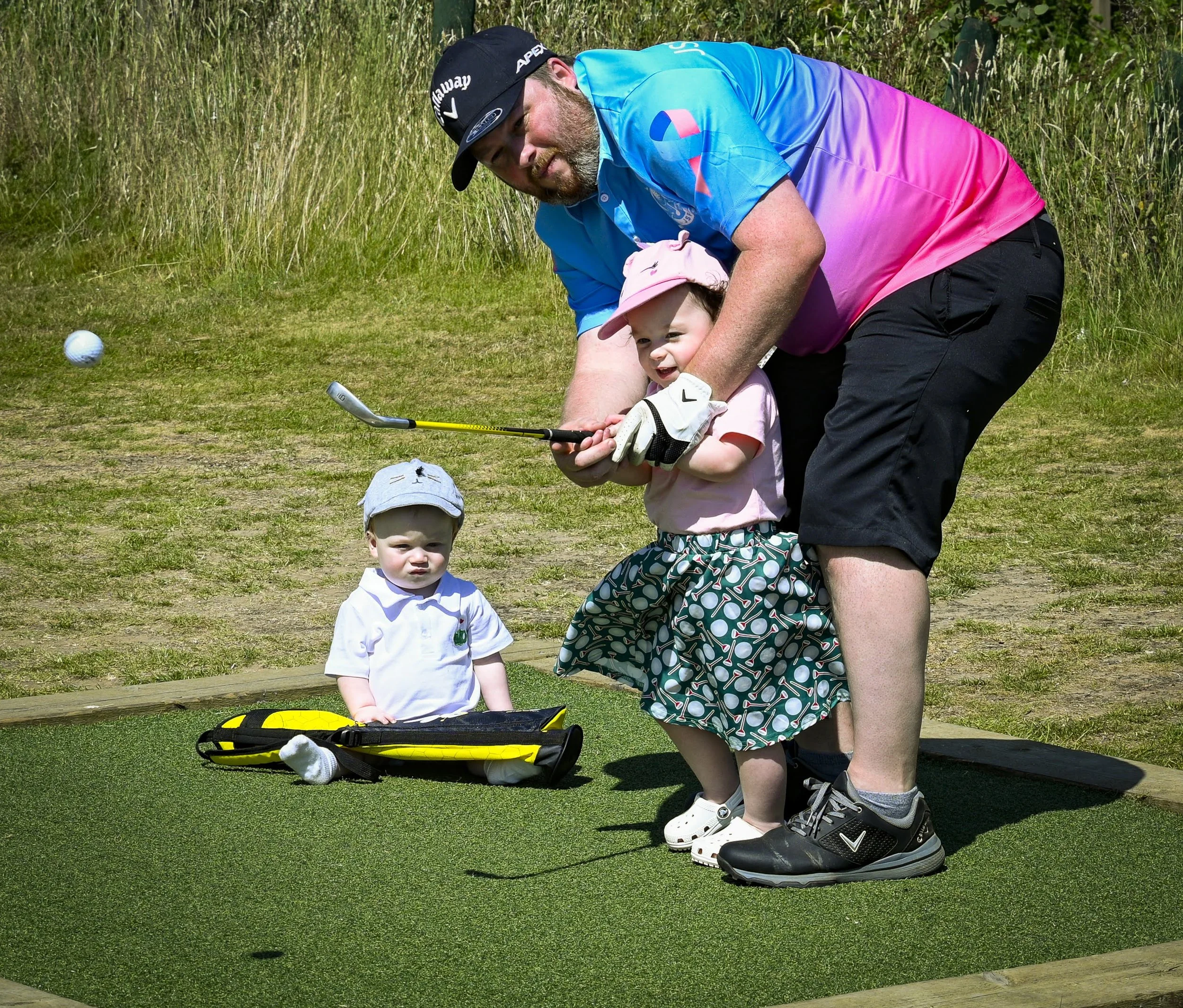 A man and a little girl are playing golf on a small green course in an outdoor setting with tall grass and trees. The man is wearing a blue and pink gradient shirt, black shorts, and a black cap, while the girl is dressed in a pink hat, a pink shirt, and patterned shorts. There are golf clubs and a golf bag nearby, and the little girl looks excited while the man is leaning over, helping her hold a golf club.