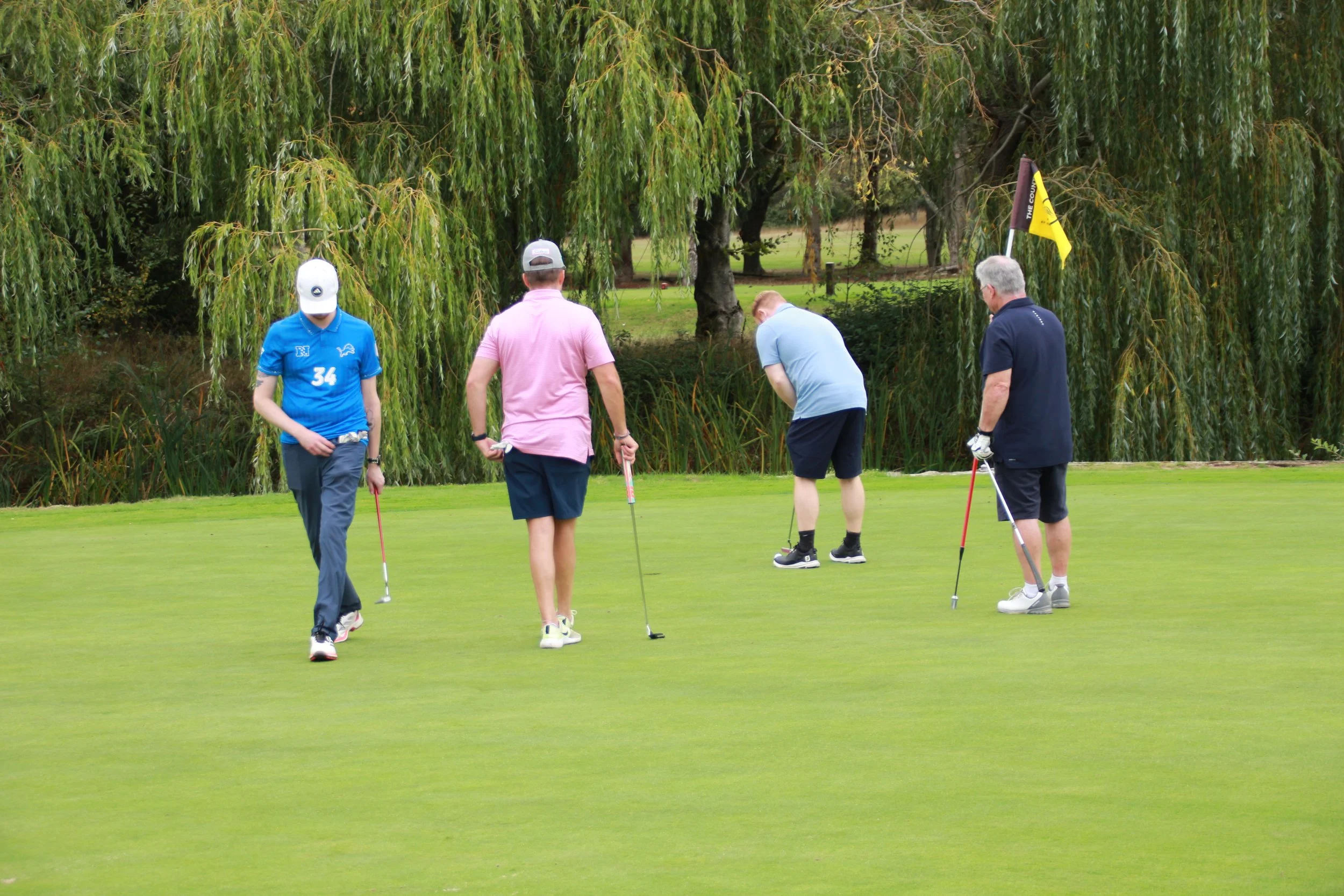 Four people playing mini-golf outdoors near tall trees, dressed casually, with a yellow and black flag on the course.