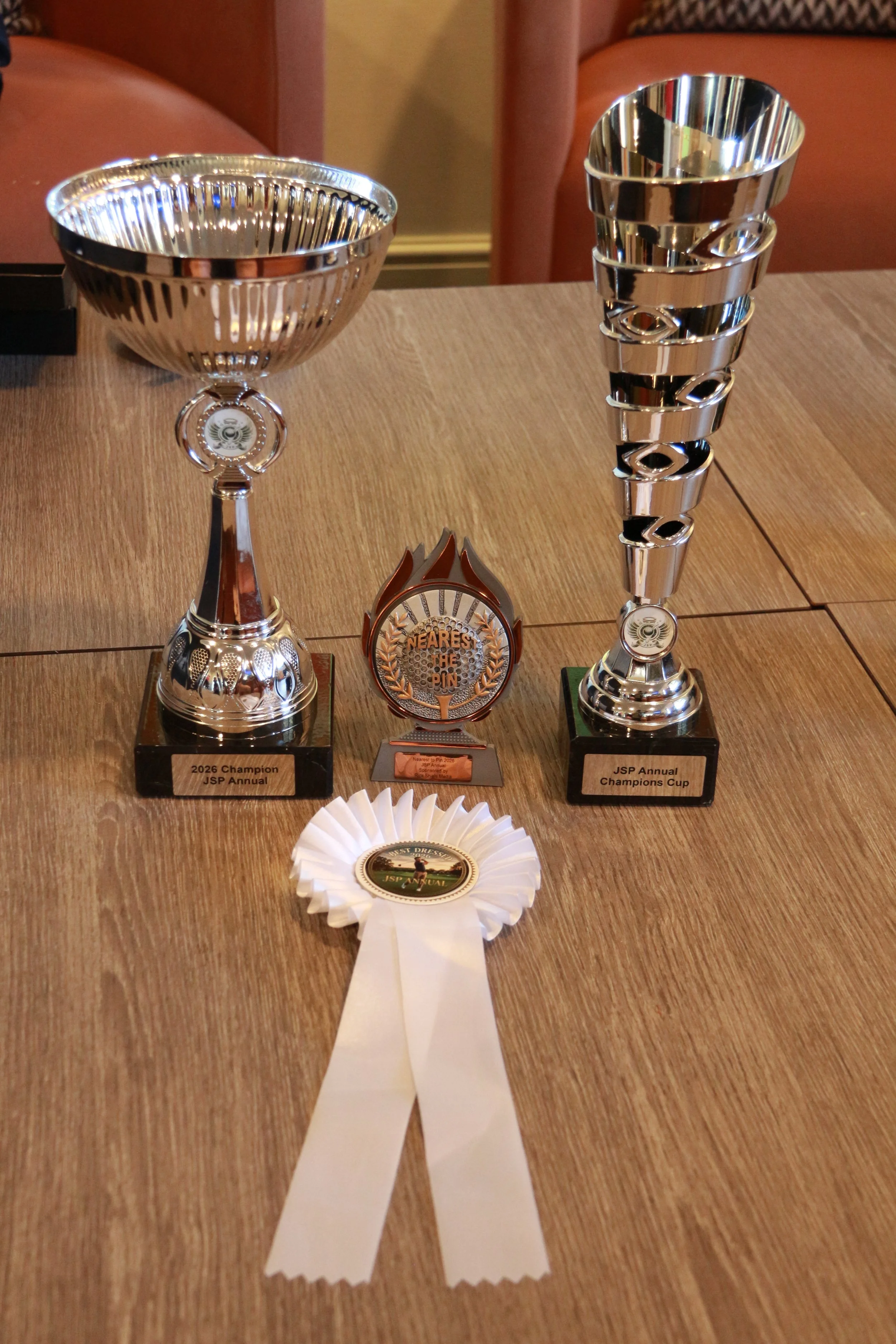 The image shows four trophies and one ribbon award displayed on a wooden table. The left trophy is labeled '2026 Champion JSP Annual,' is made of silver, and has a fluted design. The right trophy, labeled 'JSP Annual Champions Cup,' is taller with st