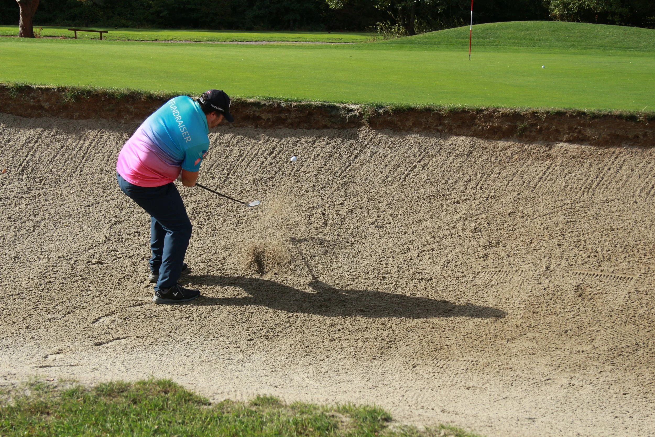 A person in a colorful shirt and black cap playing golf in a sand bunker on a golf course, with green grass, trees, and a flag in the background.