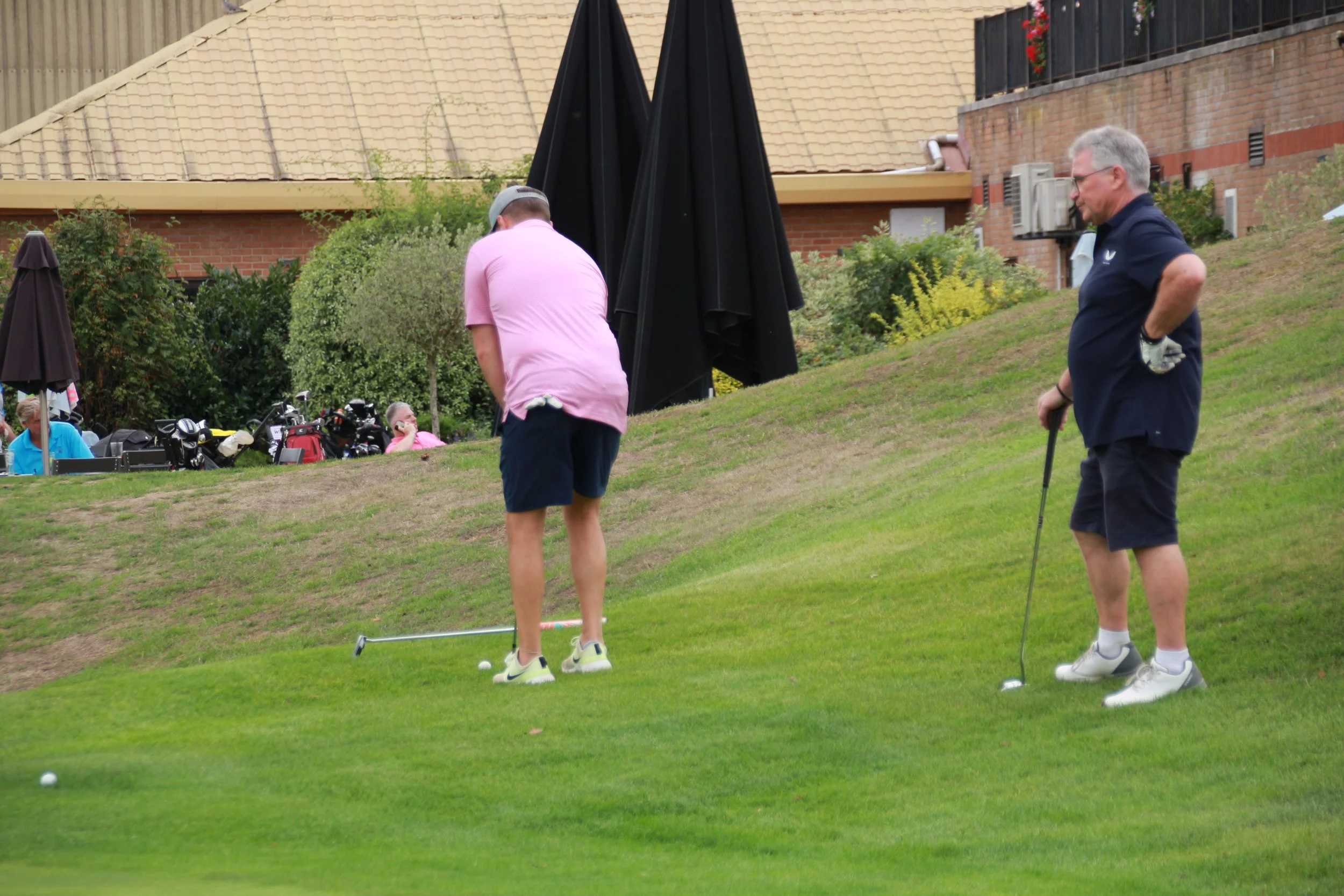 Two men playing golf on a grassy area with golf clubs and equipment nearby, one wearing a pink shirt and shorts, the other in a navy shirt and shorts, standing on a slope with a building and trees in the background.