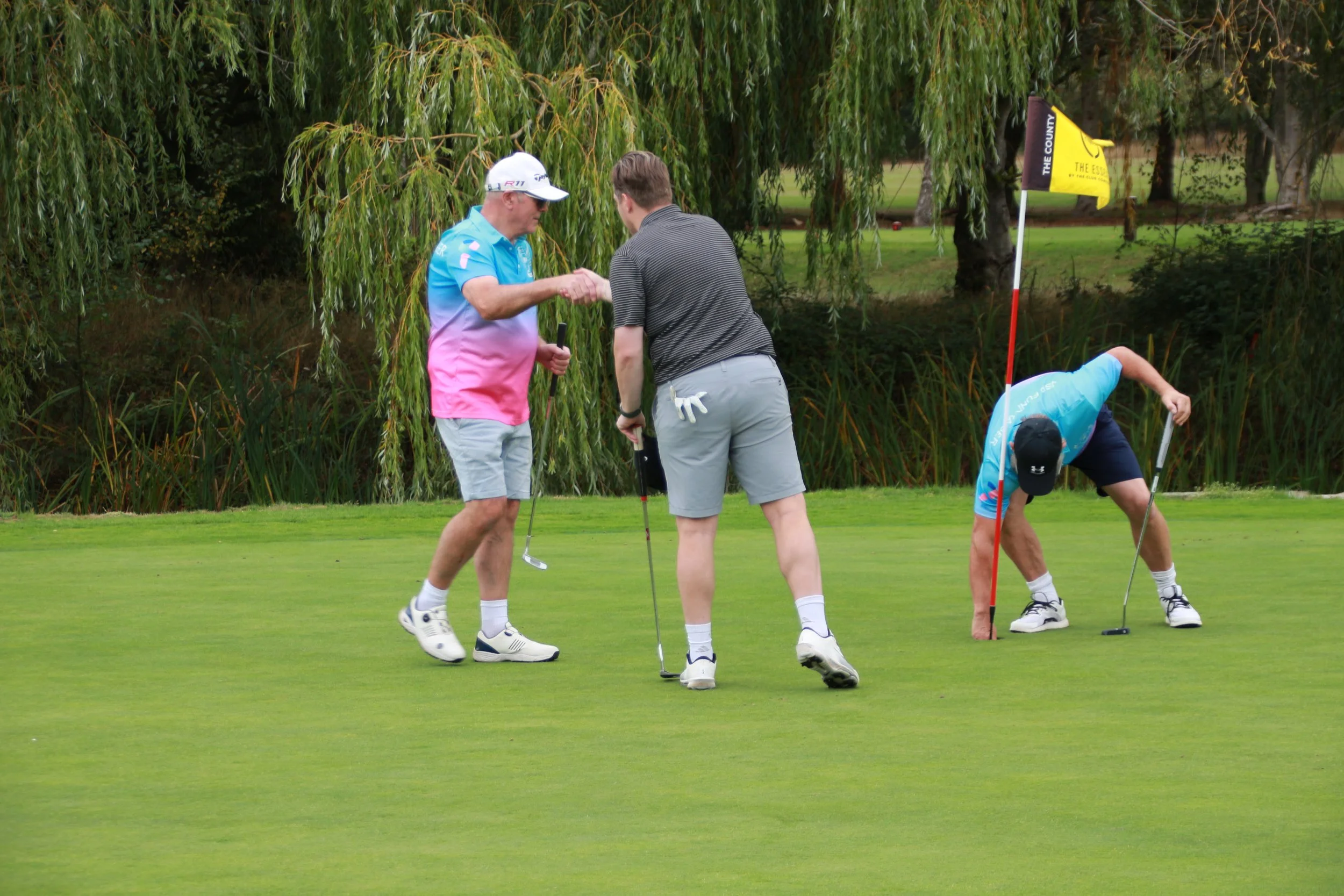 Two golfers appear to be discussing on a golf course green, with a flag and another golfer in the background.